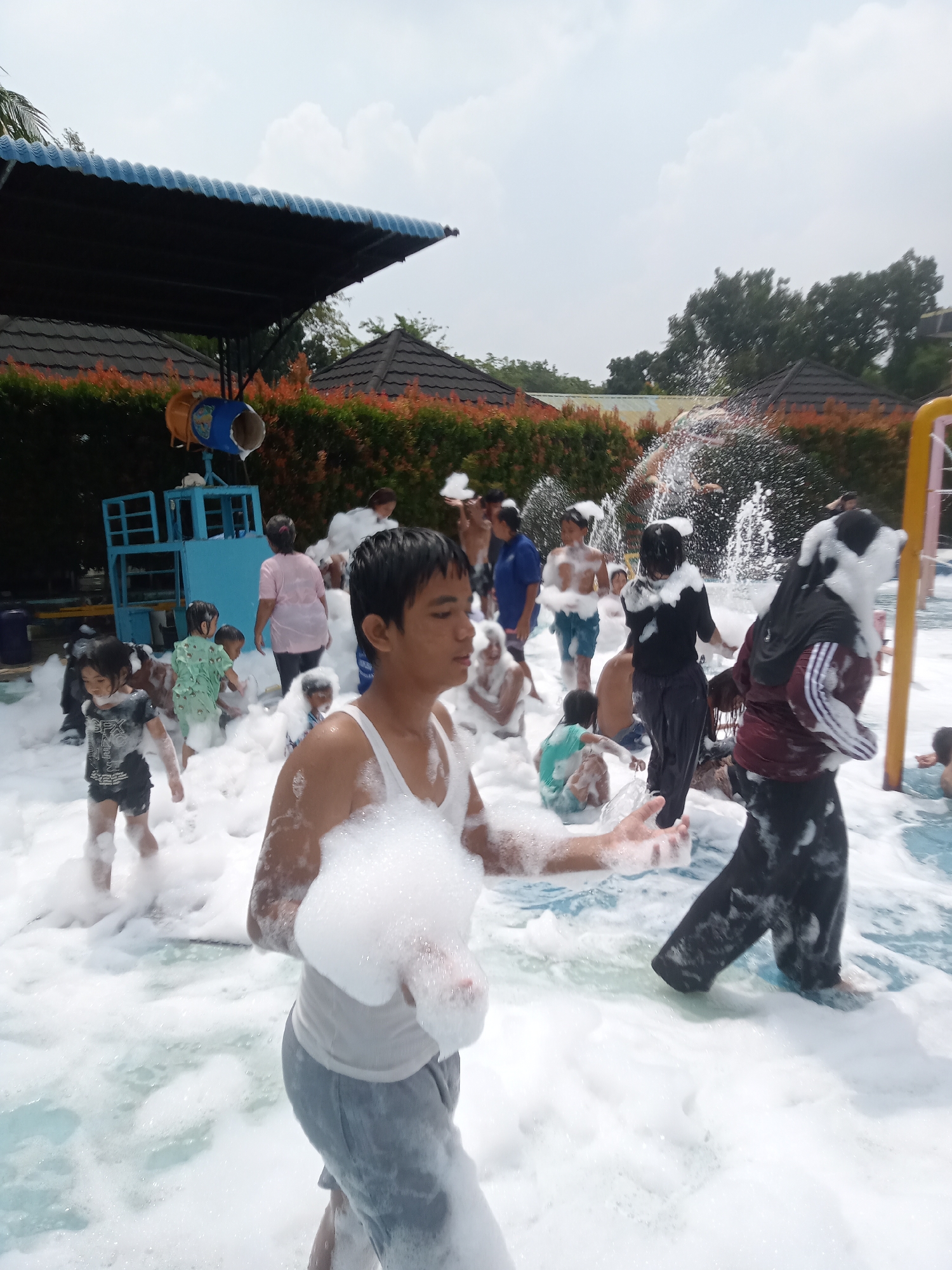 The image depicts a lively outdoor foam party in a pool, likely at a water park or resort in Baru, Indonesia. Numerous individuals, predominantly children and young adults, are immersed in a sea of white foam. Several people are actively playing and interacting within the foam, with some holding handfuls of it. In the background, a blue multi-level structure with a yellow slide can be seen, suggesting it's part of a water play area. Lush green and orange foliage borders the scene, and a dark blue awning provides shade over a section of the pool. The sky is overcast with scattered clouds.