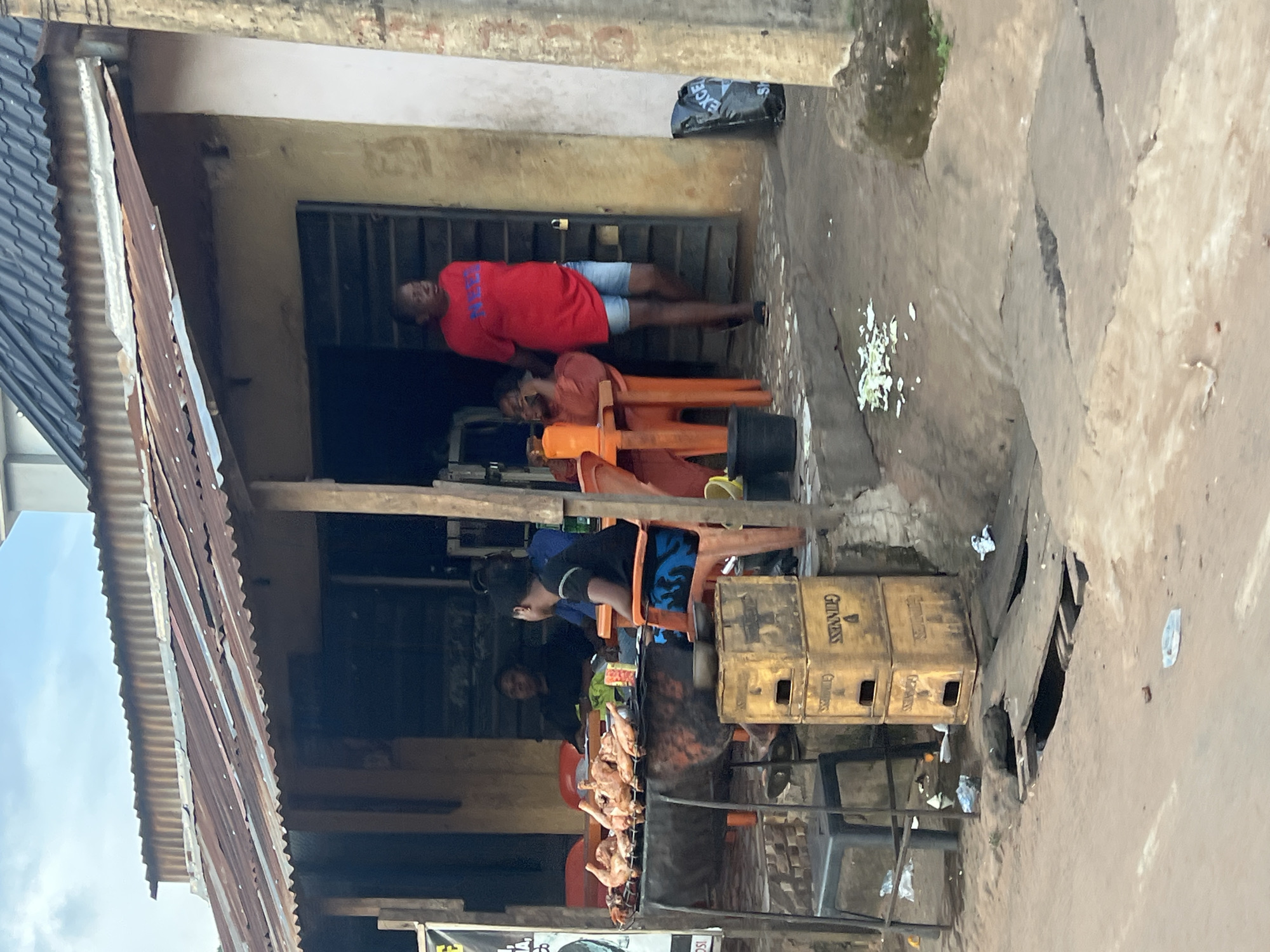 The image depicts an outdoor scene in Nkpor, Nigeria, likely in front of a business establishment. A person wearing a red t-shirt and blue shorts is lying down, seemingly resting or sleeping. Another person, wearing a black t-shirt and blue shorts, is sitting on an orange plastic chair, looking to the left. In the foreground, several whole chickens are arranged on a rack, suggesting food preparation or sale. Stacked wooden crates, some with "GUINNESS" printed on them, are visible to the right of the seated person. The setting appears to be a somewhat rustic environment with a corrugated iron roof on the left, and a rough, possibly unpaved ground. The overall impression is that of a casual, everyday scene in a Nigerian locale, possibly during daytime given the lighting.