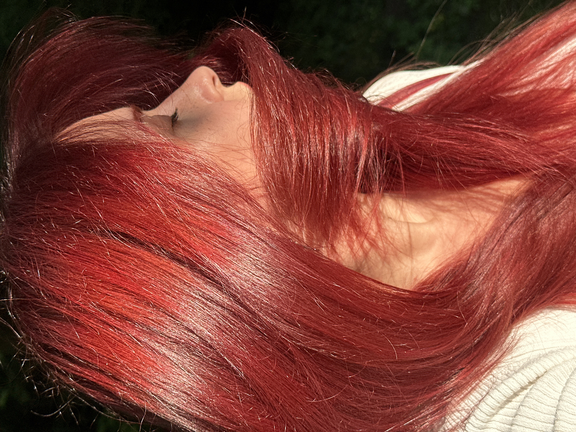 The image presents a close-up, profile view of an individual with long, vibrant red hair. The hair, illuminated by strong, directional light, cascades across the lower part of the face, partially obscuring the mouth and chin. The individual's eye is closed, revealing eyelashes, and the nose is visible in profile, showing fair skin with subtle texture. Their head is slightly tilted, suggesting a moment of repose or a dynamic pose with flowing hair. A white or off-white ribbed garment is visible on the left. The background is an out-of-focus, dark green expanse, indicating an outdoor natural setting.