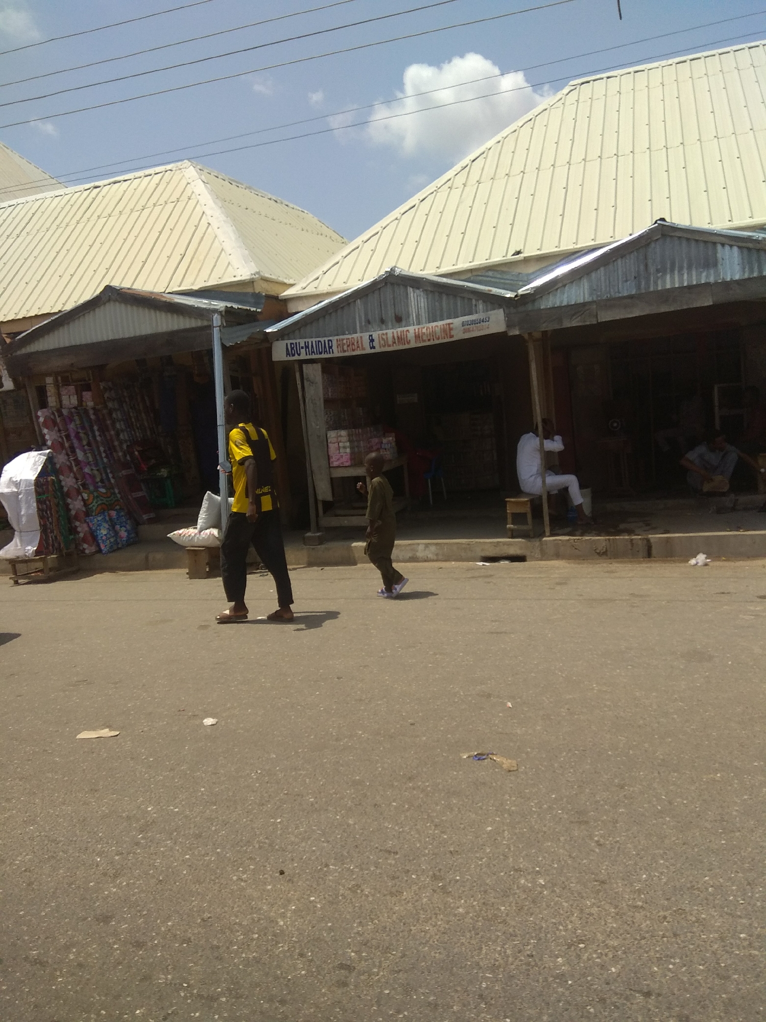 The image depicts a street scene in Jalingo, Nigeria, under a bright, partly cloudy sky. In the foreground, a paved road shows signs of wear and scattered debris. A young boy in a green outfit walks across the road from right to left, his face obscured by the angle. Behind him, a man in a yellow shirt with black detailing and black pants walks towards the left. He is wearing sandals.
The background features a row of shops with corrugated metal roofs. One shop prominently displays a sign that reads "ABU-HAIDAR HERBAL & ISLAMIC MEDICINE" with a phone number. In front of this shop, a man dressed in white is seated on a stool, facing away from the camera. Further to the right, another individual is crouched down, appearing to be working on something. To the left of the "ABU-HAIDAR" shop, stacks of colorful textiles are displayed outside another establishment. A white tarp-covered object is also visible in front of this shop. Power lines crisscross the sky above the rooftops.
