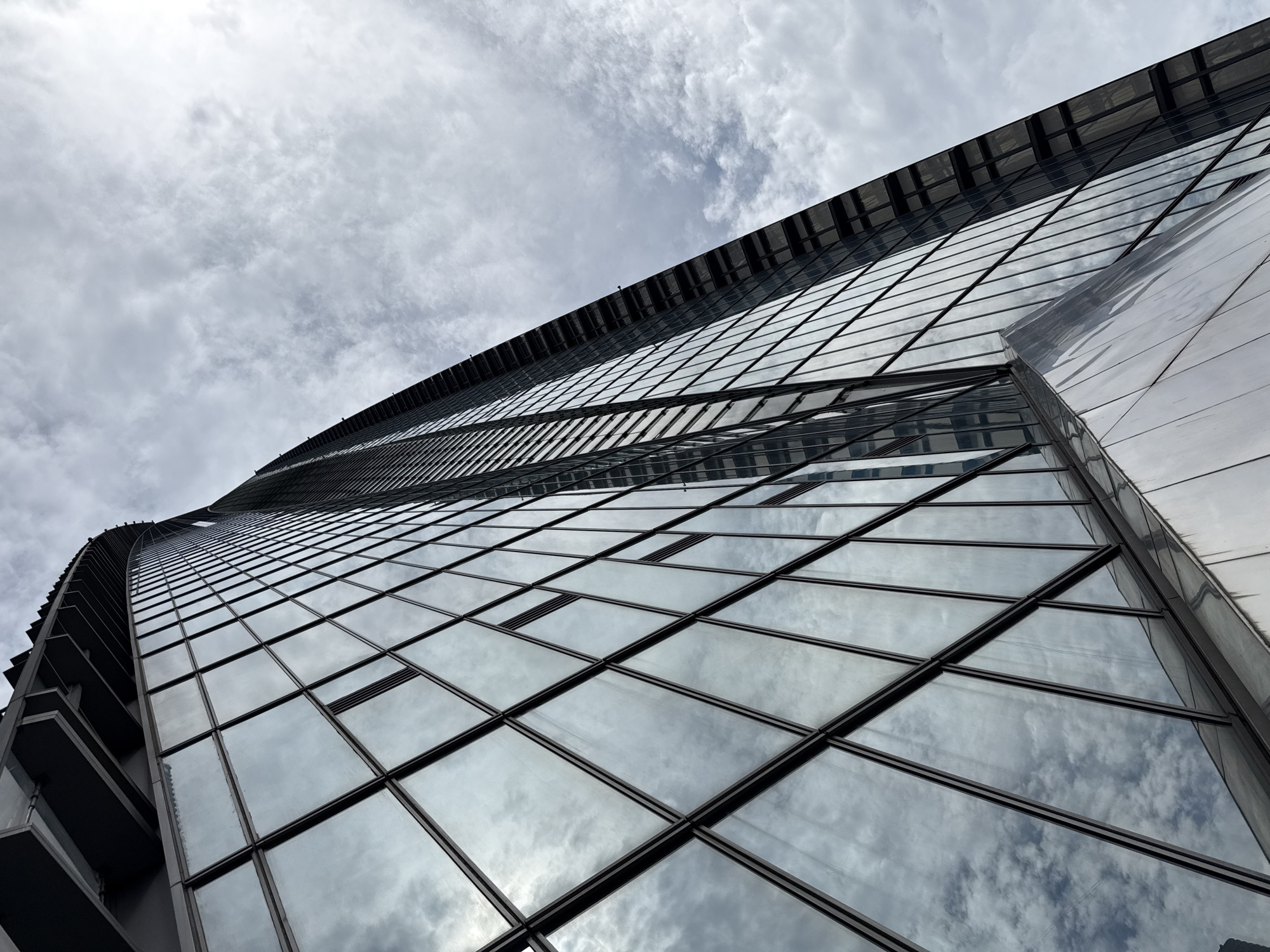 An upward-angle, low-angle shot captures the exterior of a modern skyscraper in Tianhe District, China. The building features a distinctive curved facade covered in reflective glass panels, which mirror the cloudy sky above. The geometric patterns of the windows and their dark frames create a repetitive, linear design. The upper portion of the skyscraper shows a darker, more angular structure. The scene is devoid of any people or vehicles, focusing solely on the architectural details of the building and the atmospheric conditions. The lighting is overcast, contributing to a muted and somewhat dramatic mood.