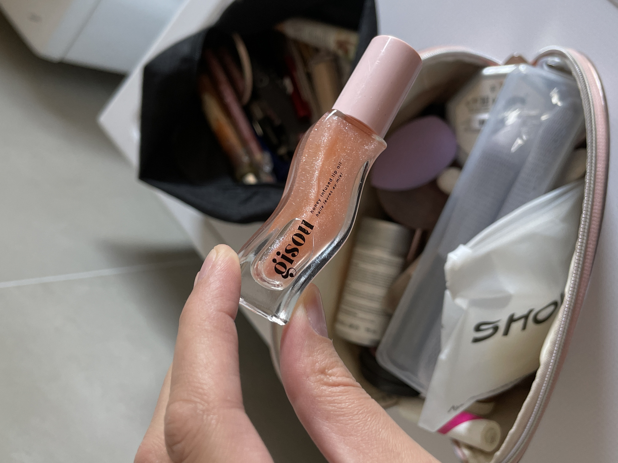 A hand holds a bottle of Gisou honey-infused lip oil in front of an open makeup bag, revealing various cosmetic items inside. The bag is set near what appears to be a container or box. The location, Подшивалово, Russia, cannot be confirmed from the image.