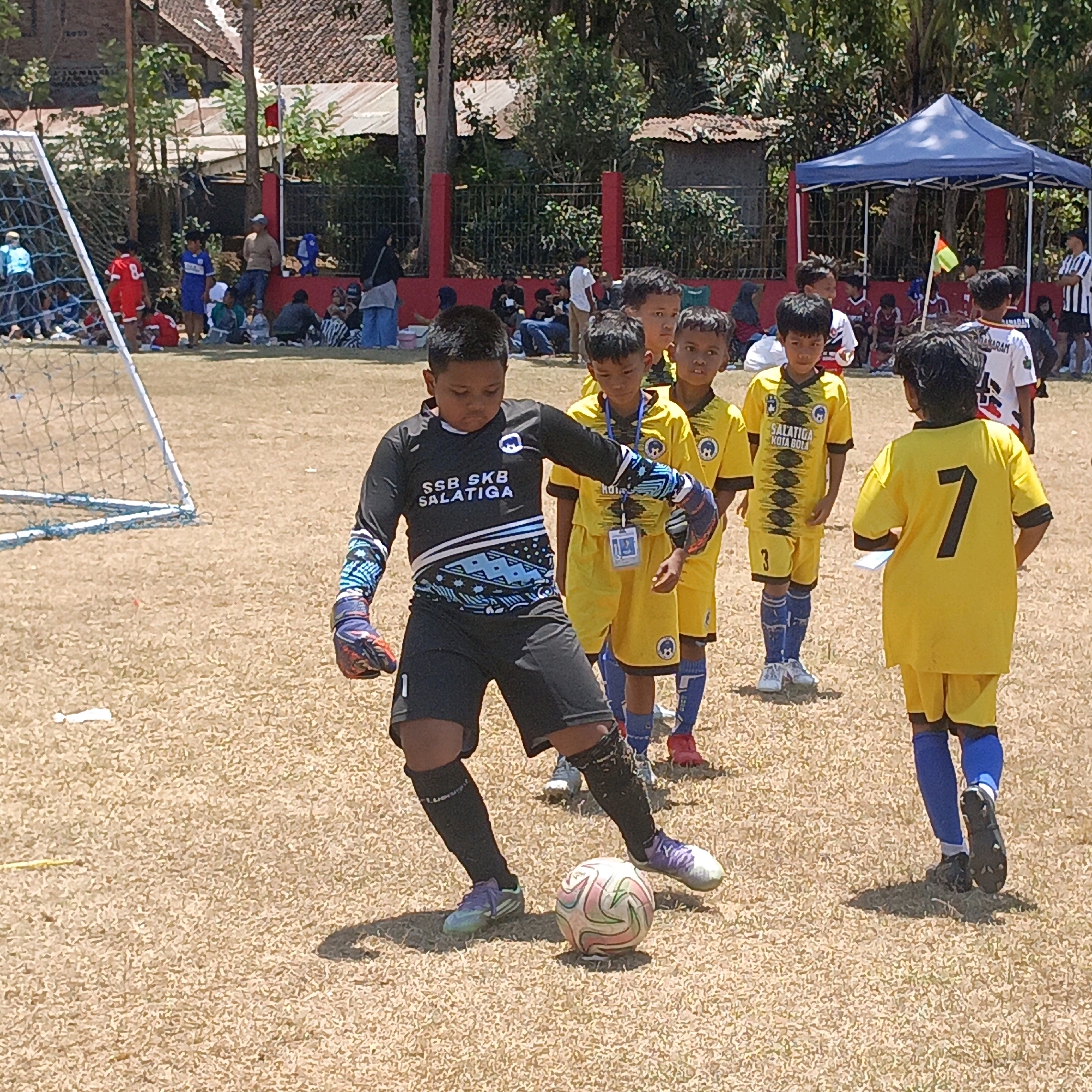 A group of young boys are playing soccer on a dry, grassy field in Krajan Lor, Indonesia. The main focus is on a boy in a black goalkeeper uniform, wearing gloves and black shorts, actively engaged with a soccer ball at his feet. Behind him, several other boys in yellow soccer uniforms are also present on the field. In the background, spectators are visible, some seated under a blue canopy. The environment appears to be a community sports event, with trees and buildings visible in the distance.
