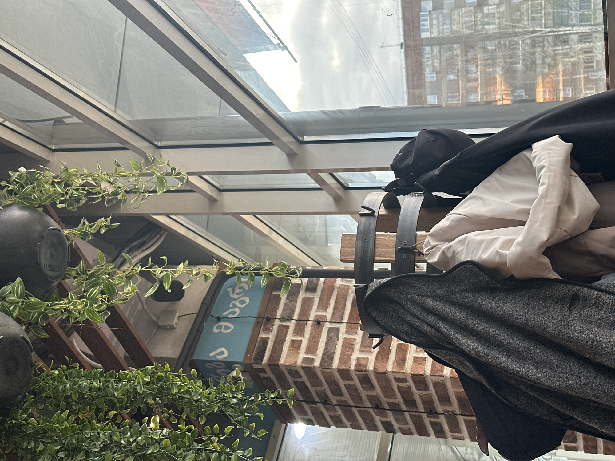 The image shows an indoor setting with a glass ceiling and a view of a building across the street, suggesting an urban location. A rack made of a wooden plank and metal straps holds several garments: a black baseball cap, a white garment (possibly a shirt or jacket), and a gray tweed coat. A potted plant with green and white variegated leaves hangs nearby. The background includes a brick structure with some blue signage. The scene has a casual, slightly cluttered feel, suggesting a restaurant or shop interior. No people are visible.