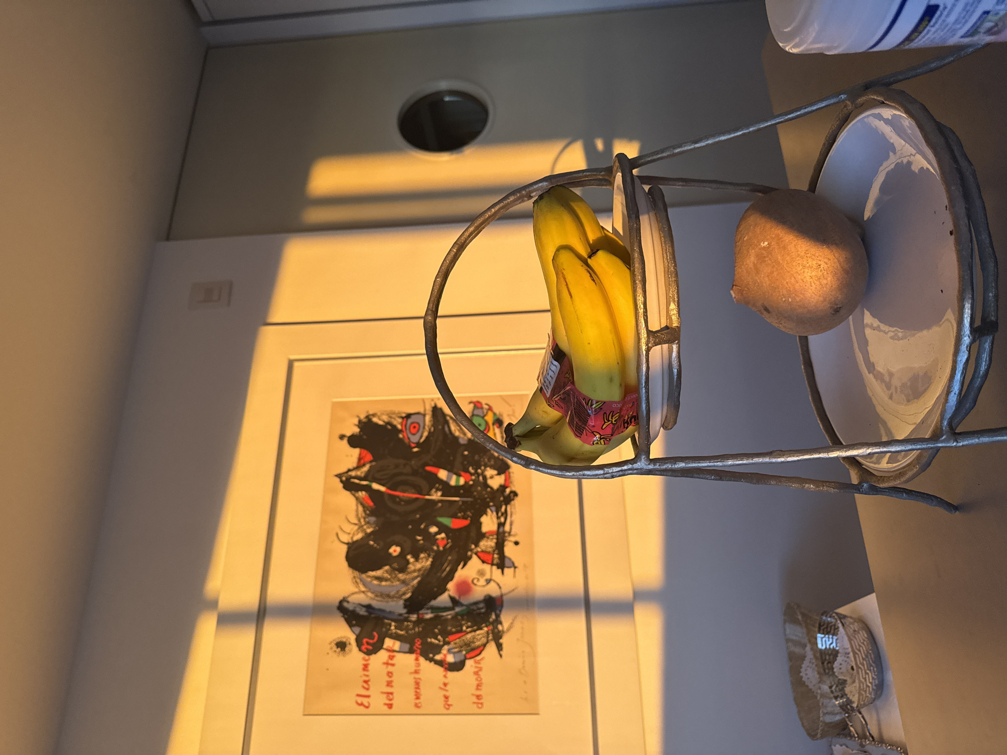 The image shows a still life arrangement in an indoor setting. A metal fruit basket holds a bunch of bananas and a single kiwi. The background includes a framed abstract print on a wall and a ceiling fixture with a circular opening. The lighting appears to be warm, possibly from late afternoon sunlight. The objects are positioned on a light-colored surface, perhaps a countertop. The precise location within Mexico City, Mexico, cannot be determined from this image alone.