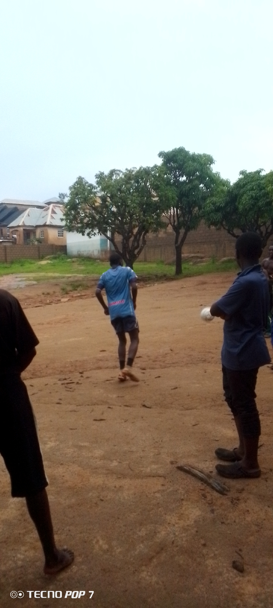 In an outdoor scene of Unknown, Unknown, three individuals are present. The person furthest to the left, partially visible, is wearing dark clothing. In the center, a person in a blue t-shirt with a design on the back and dark shorts is kicking a light-colored ball with their right foot, suggesting they are playing soccer. To the right, a person in a dark blue t-shirt and dark pants is facing away from the viewer, holding a white object, possibly another ball, in their hands. The environment consists of a dirt or sand-covered ground with scattered debris. In the background, several trees with green foliage are visible, and behind the trees are buildings with pitched roofs, indicative of a residential area. The sky is overcast and light in color.