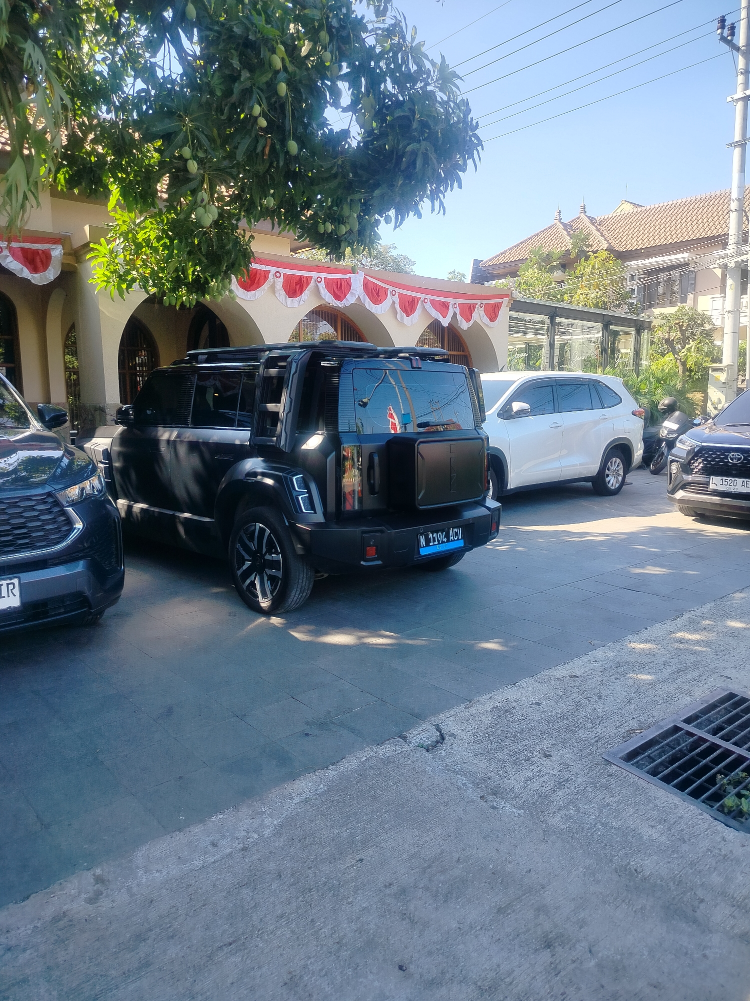In Surabaya, Indonesia, a black SUV, possibly a Wuling Almaz, is parked on a paved surface. The SUV has a license plate that reads "N 1194 ACV". To the left of the black SUV, another vehicle, a dark-colored SUV, is partially visible, with its front bumper and a portion of its license plate, which starts with "LR", showing. To the right of the black SUV, a white SUV, likely a Honda BR-V, is parked, and further to the right, a dark motorcycle is visible. Behind the vehicles, a building with arched entrances and white and red festive decorations is present. Overhead, a tree with green mangoes hangs its branches. The sky is clear and blue, and power lines are visible. The ground is paved with concrete tiles and some areas are textured concrete. A metal grate, likely a drain cover, is located in the foreground. The scene appears to be a street or a parking area outside a building.