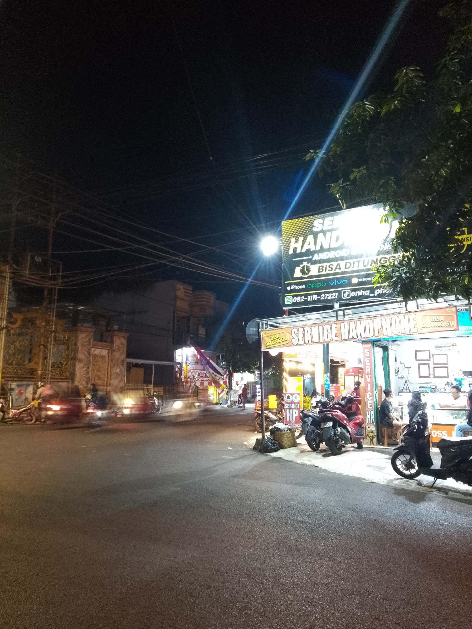 The image depicts a street scene at night in Lamongan, Indonesia, featuring a mobile phone service shop. A prominent yellow and black sign above the shop reads "SERVICE HANDPHONE" and "BISA DITUNGGU" (meaning "can be waited for"). Below this, smaller text indicates support for various brands like iPhone, Oppo, Vivo, and Xiaomi, along with a phone number and social media handle.
In front of the shop, several motorcycles are parked and visible. The shop itself appears to be an open-air establishment with a counter where staff and customers are interacting. Customers are seated on stools, and one person is standing behind the counter. The environment suggests a busy commercial area with streetlights and building lights illuminating the scene.
The street in the foreground shows motion blur from passing vehicles, indicating traffic. A building with ornate yellow gates is visible to the left, possibly a temple or a traditional building. Power lines are strung across the dark sky. The overall impression is a snapshot of nighttime commerce and street life in an Indonesian city.