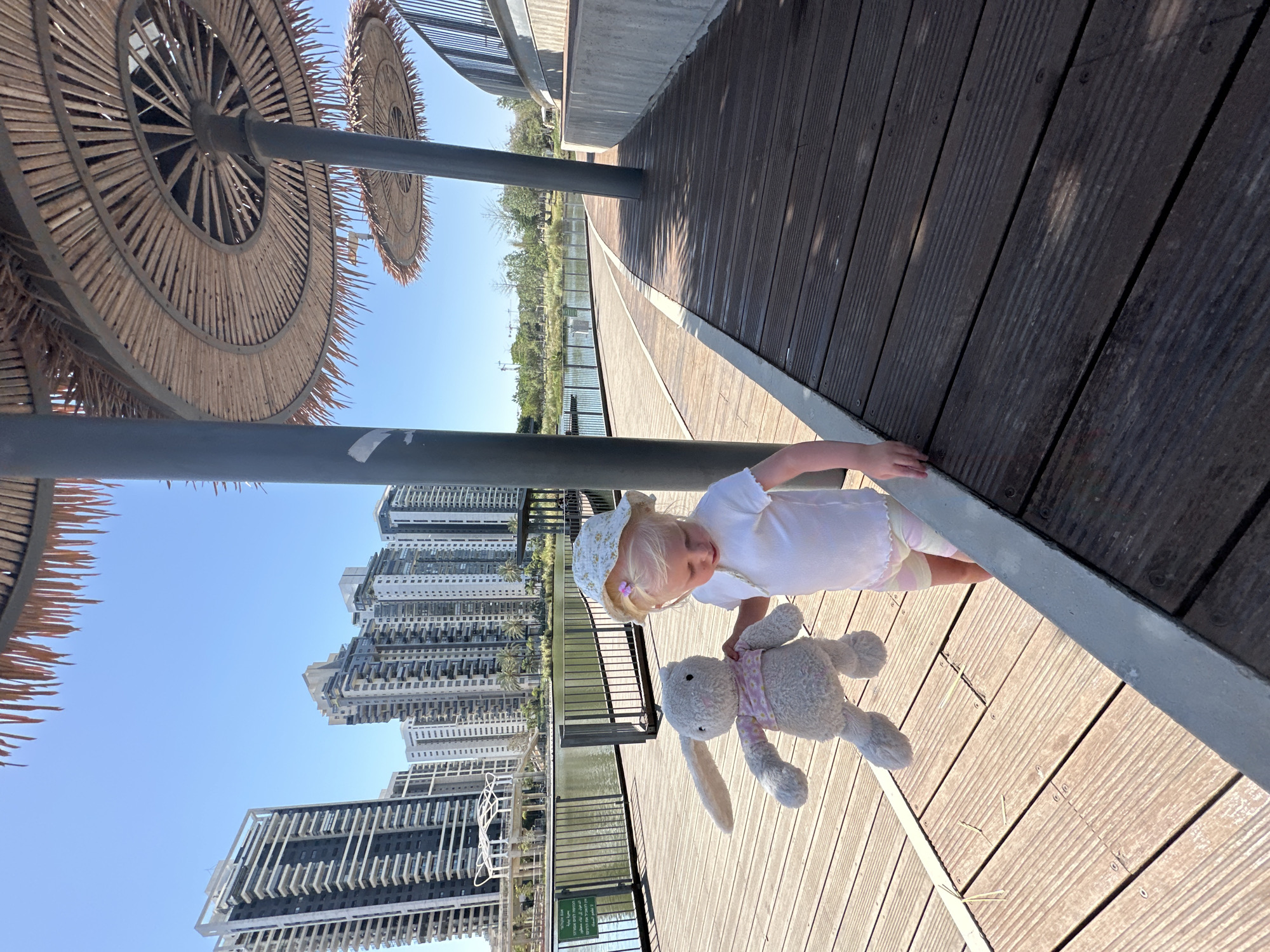The image depicts a young Caucasian child holding a stuffed rabbit on a wooden boardwalk beside a body of water in Petah Tikva, Israel.
The child, appearing to be a toddler or preschooler, with blonde hair and fair skin, stands towards the right side of the frame. She wears a white ribbed short-sleeved top, light-colored shorts with horizontal pink and white stripes, and a light-colored bucket hat with a subtle floral or patterned design. A small purple hair clip is visible on the left side of her head, underneath the hat. Her gaze is directed downwards towards the wooden surface and a grey concrete edge. In her left arm, she holds a grey stuffed rabbit, which has long ears and wears a light pink patterned shirt. The rabbit's body faces the viewer.
The child is positioned on a light brown wooden boardwalk composed of horizontal planks. To her immediate right, a darker brown wooden deck runs parallel, separated by a grey concrete curb. Several dark grey metal pillars rise vertically from the boardwalk, supporting large, circular shade structures made of natural, woven, straw-like material arranged in concentric patterns. At least three such structures are visible, two prominently overhead and to the right, and one partially at the top edge of the frame.
To the left of the child and the pillars, a calm body of water, likely a lake or pond, extends into the midground. A dark metal fence or railing separates the boardwalk from the water. A white informational sign with Hebrew text is visible on the left side of the boardwalk, near the water's edge.
In the background, a dense cluster of numerous modern high-rise apartment buildings dominates the skyline beyond the water. These buildings feature contemporary architectural designs with various facades in shades of dark grey, white, and light grey, and multiple balconies. Green vegetation, including trees and shrubs, lines the far bank of the water, separating it from the buildings. The sky above is clear and bright blue. In the distant background, on the far left, what appear to be construction cranes are faintly visible.
The specific location is an urban park or promenade in Petah Tikva, Israel, characterized by its modern residential developments surrounding a recreational lake, consistent with areas such as the Petah Tikva Lake Park.