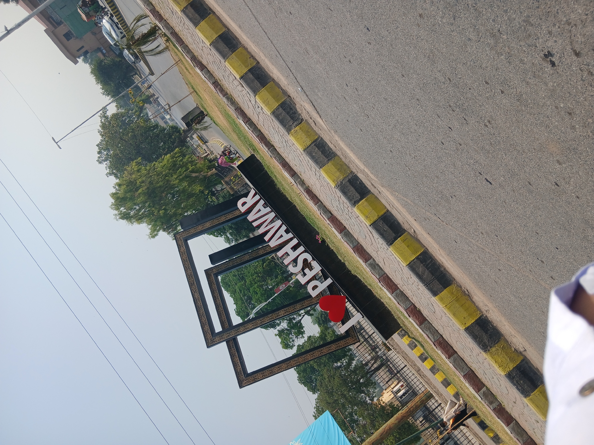 An outdoor, medium shot captures a tilted view of a large, illuminated sign that reads "I ❤️ PESHAWAR" in white, three-dimensional letters. The sign is set against a backdrop of trees and a pale sky. The text is partially obscured by a decorative black and gold frame structure. In front of the sign, a red heart symbol is visible. The sign is situated on a paved area with a border of yellow and black checkered tiles. In the background, a street with cars and a few pedestrians is visible, suggesting an urban environment. The overall lighting indicates it is daytime, with a bright, slightly hazy sky. The image appears to be taken from a street-level perspective, possibly from a moving vehicle given the tilt.