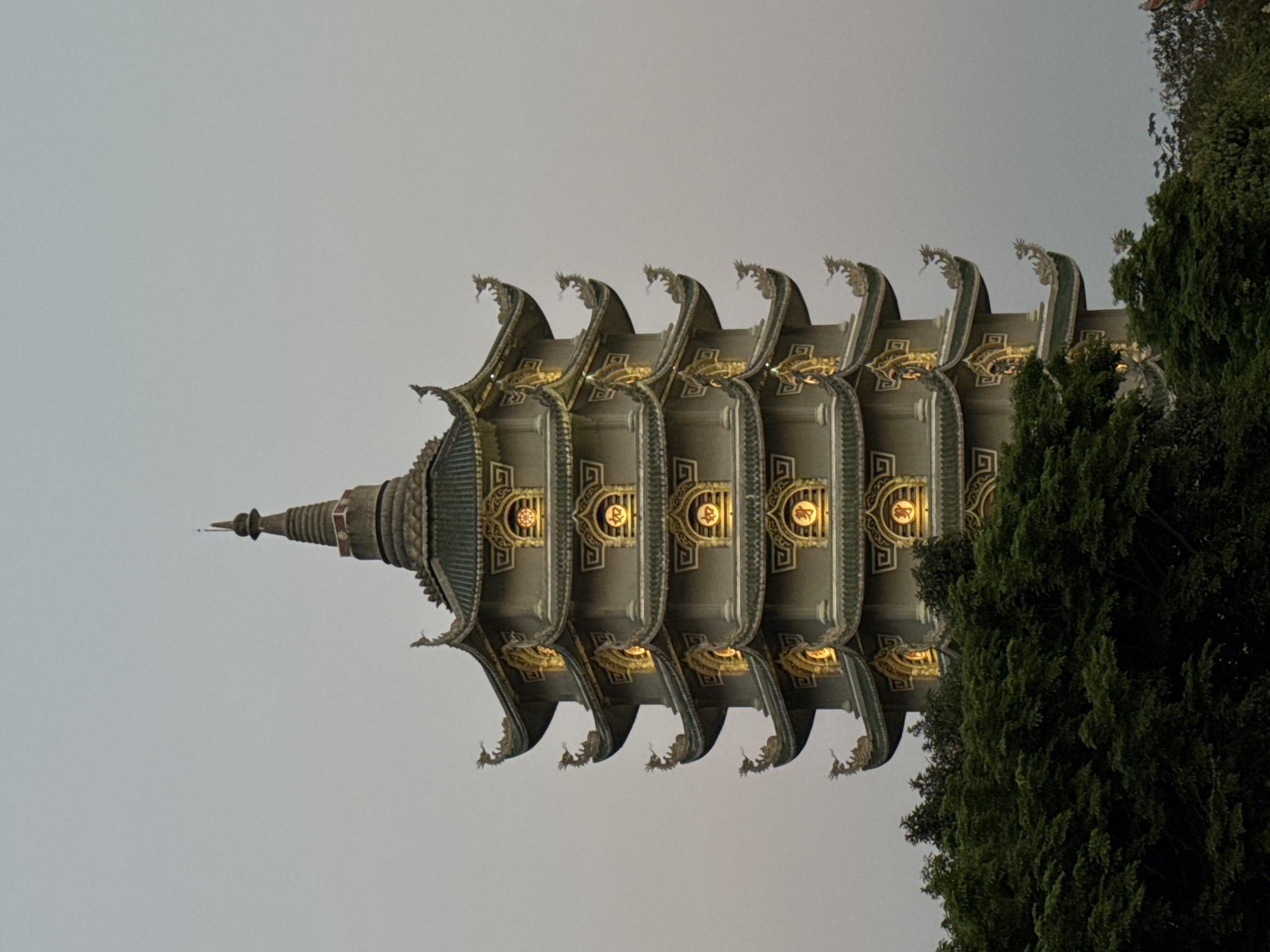 A low-angle, horizontal outdoor shot depicts a multi-tiered pagoda against a gray sky. The pagoda is composed of stacked, gray stories featuring upturned eaves adorned with dragon-like sculptures. Each story has a geometric design and an illuminated circular emblem. To the right of the pagoda, lush green trees are partially visible. The location is likely in the city of Đà Nẵng, Vietnam.