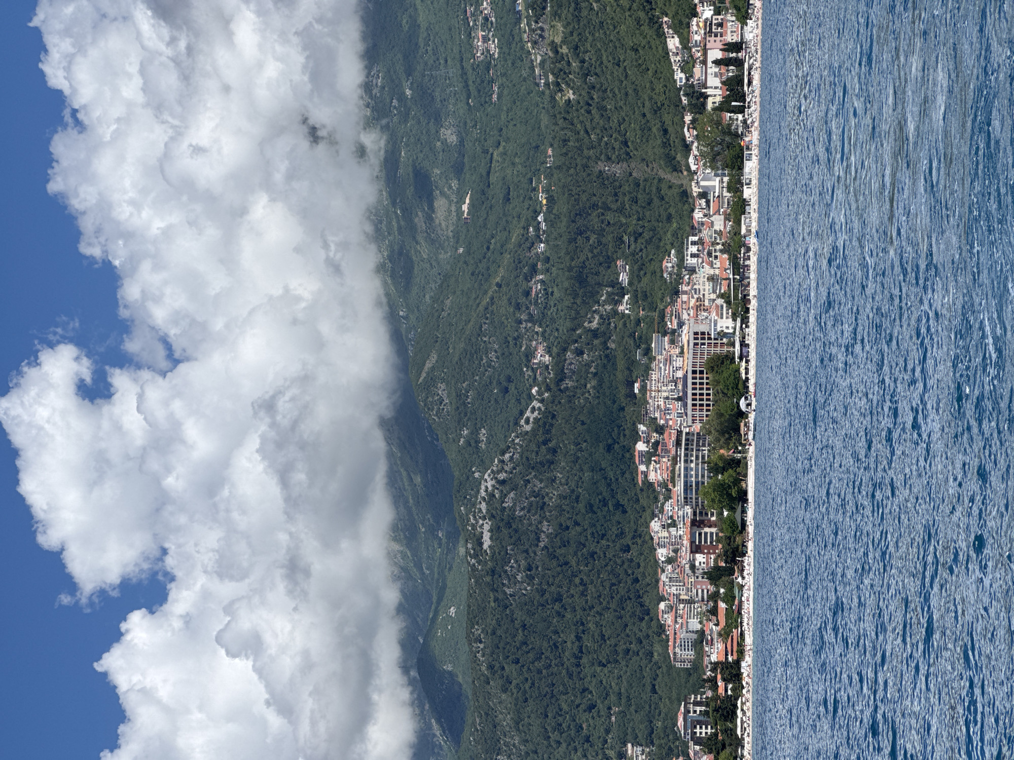 The image displays a panoramic coastal scene in Budva, Montenegro, viewed from an elevated perspective. The scene is bathed in bright daylight under a partially cloudy sky.
On the left side of the frame, a vibrant blue sky is visible, adorned with large, fluffy white cumulus clouds that stretch across a significant portion of the upper left and central sky. Below the sky and clouds, majestic green mountains dominate the mid-ground. These mountains are densely covered with lush forestation, and some rocky outcrops are discernible, indicating rugged terrain. The mountain slopes extend down towards the coastline.
At the foot of the mountains and hugging the shoreline is a bustling town. It features a dense cluster of buildings, characterized by a mix of architectural styles. Many structures have light-colored facades and distinctive red-tiled roofs, common in Mediterranean regions. There are also taller, more modern multi-story buildings intermingled with what appear to be older, more traditional residences. The town stretches along the coast, indicating a significant settlement or resort area.
To the right, a wide expanse of deep blue water, likely the Adriatic Sea, fills the remaining portion of the frame. The surface of the sea is textured with numerous small ripples and glints, reflecting the bright sunlight, suggesting gentle waves or currents. While specific details are too small to discern clearly, the presence of the town directly alongside the water implies a popular coastal destination, possibly with a beach or promenade area.
The time of day appears to be mid-day, given the strong, clear light. The weather is sunny with scattered clouds, creating a pleasant atmosphere. No discernible text is visible within the image.