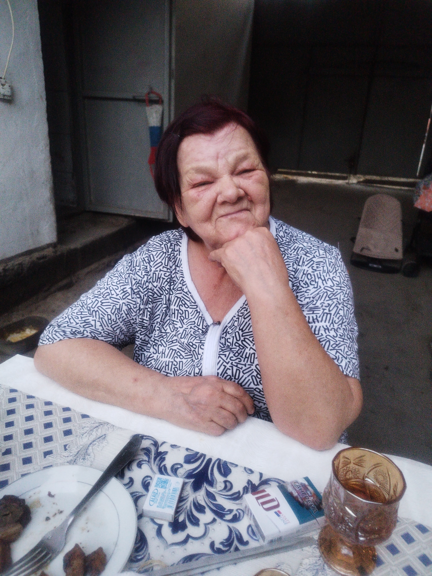 An elderly woman with short, dark red hair and fair skin is seated at a table outdoors. She is wearing a black and white patterned shirt with a V-neck and a zipper. Her right hand is resting on her chin, and her left arm is bent at the elbow and resting on the table. She has a slight smile on her face.
The table is covered with a white tablecloth, and on it are a plate with leftover food, a fork, a packet of cigarettes, and a glass filled with a brown liquid. The background appears to be an outdoor setting with a concrete wall to the left and a dark, indistinct area to the right, possibly a building or an awning. A fire extinguisher with red and white bands is visible on the wall in the background. The scene suggests a casual outdoor meal or gathering. The specific location within Shymkent, Kazakhstan cannot be determined from the visual information alone.