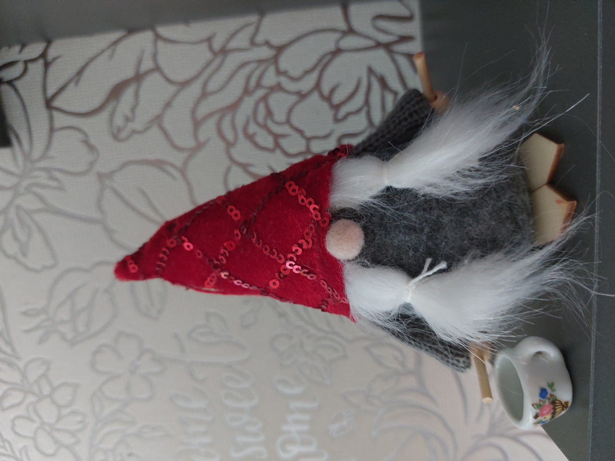 This close-up image displays a festive gnome figurine, positioned sideways on a patterned surface. The gnome is characterized by its pointed red hat, which is covered in small, shiny red sequins. A small, light pink, round nose protrudes from beneath the hat. The gnome's body is made of a grey, knitted fabric, and two white, fluffy pigtails, tied with thin white string, extend from under the hat, resembling hair or a beard.
To the lower right of the gnome, a miniature white ceramic mug is visible, adorned with small blue and pink floral patterns and green leaves. Behind the gnome, several small, light brown rectangular blocks are present.
The gnome rests on a light-colored surface featuring a subtle, raised floral pattern in a lighter shade. Faint, decorative script text is partially visible on this surface, appearing to spell out words such as "Sweet" and possibly "Home," though much of it is obscured and out of focus. The background above this patterned surface is a dark, uniform grey or black. The image captures a static display; no activity is taking place. The scene appears to be an indoor setting with even lighting. While the location context is Chaikovsky, Russia, no specific visual elements within the image directly indicate this.