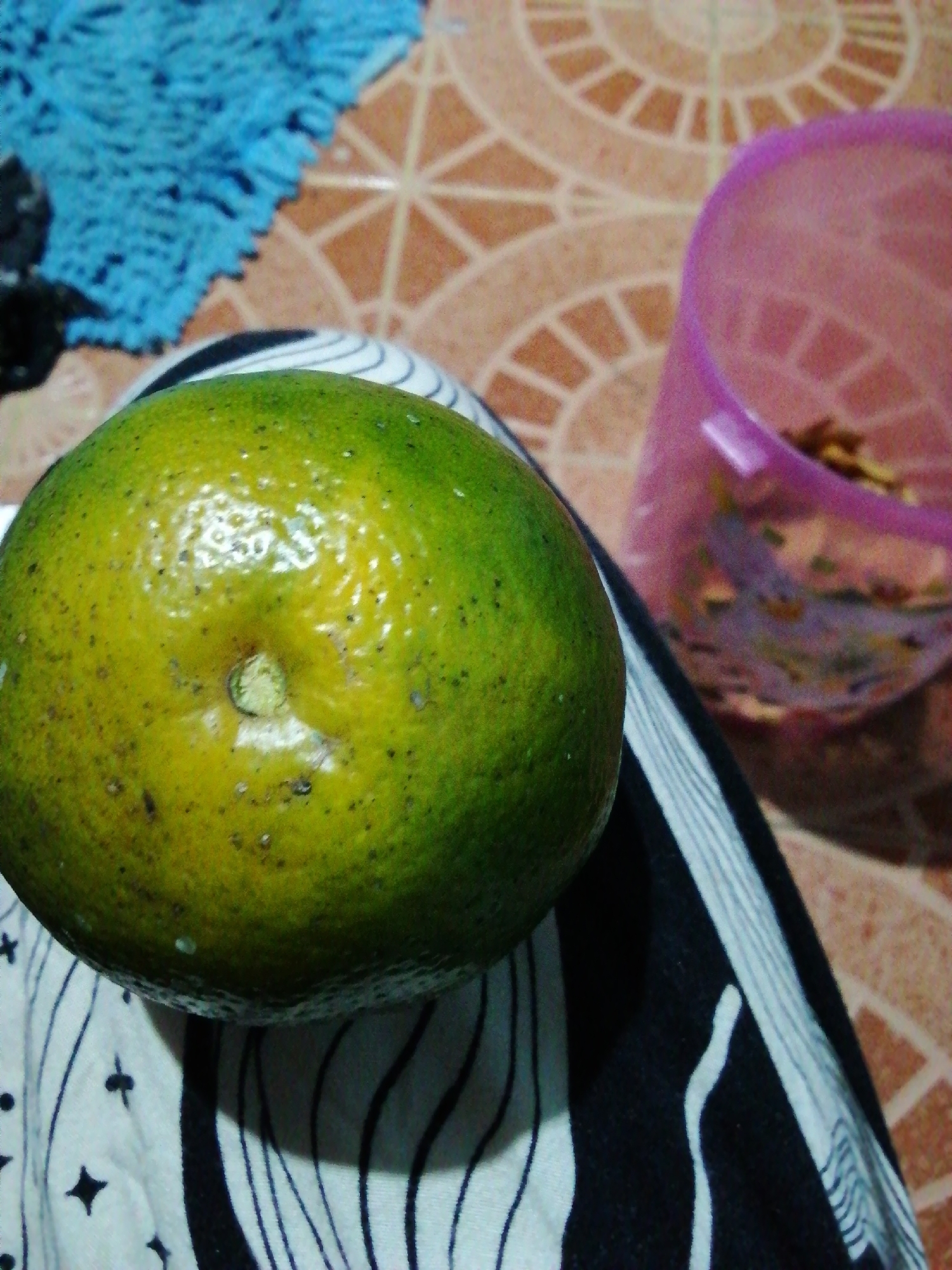 A close-up, slightly blurry, overhead shot shows a green and yellow citrus fruit, likely an orange or lime, positioned on a black and white striped cloth. The fruit is the primary subject, filling much of the left side of the frame. In the background, to the right of the fruit, a translucent pink plastic container is visible, partially filled with what appears to be a light-colored snack or food item. The surface beneath the cloth and container is a tiled floor with a geometric pattern in shades of orange and cream. To the upper left, a blurred patch of a bright blue, fuzzy object, possibly a rug or mat, is visible. The lighting is dim, with reflections on the fruit suggesting a light source from above. The scene is indoors, in what appears to be a domestic setting.