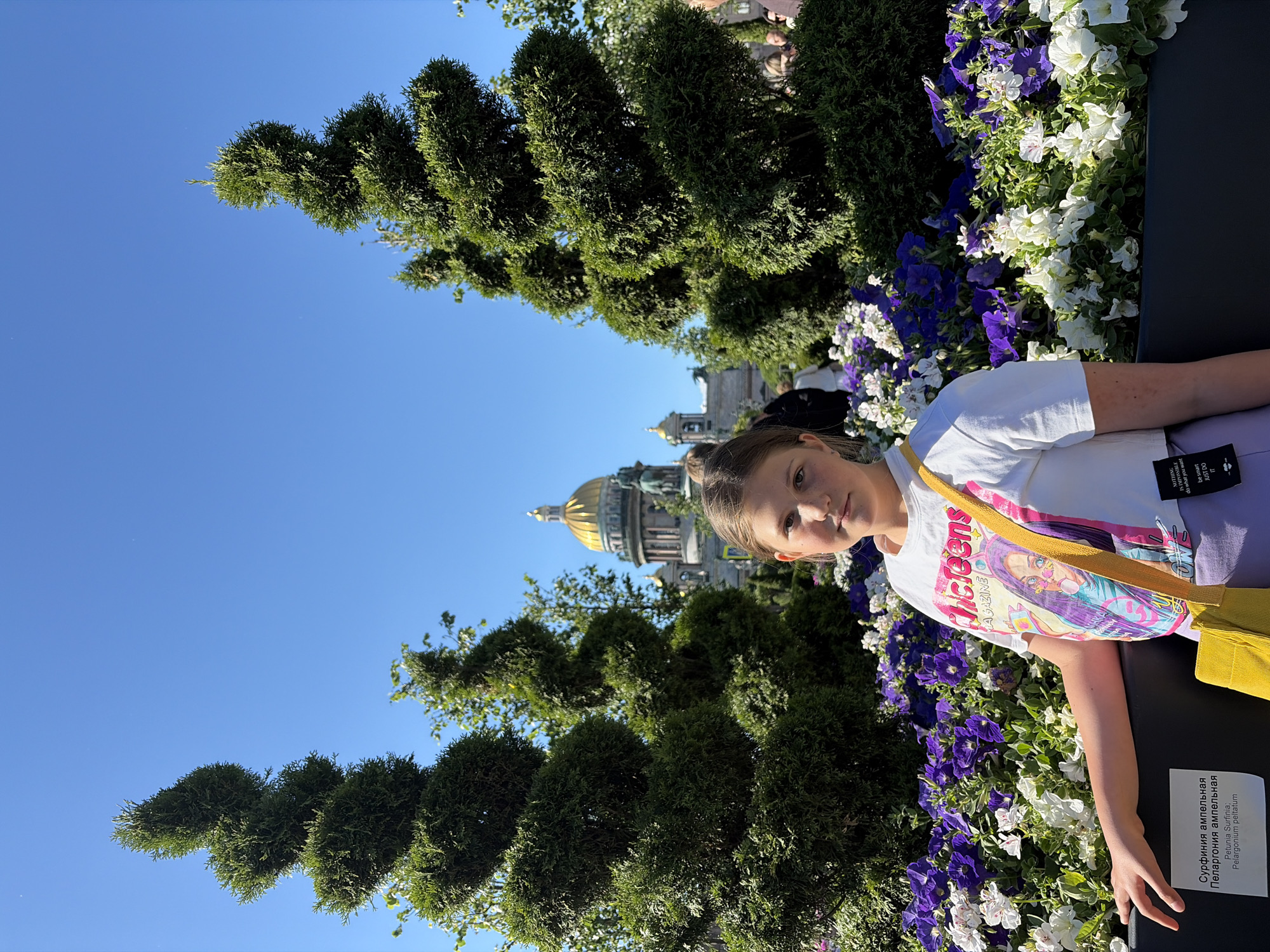 A young girl stands in front of a flower bed with purple and white petunias. Behind her, two spiral-shaped evergreen trees frame a view of St. Isaac's Cathedral in Saint Petersburg, Russia, identifiable by its golden dome and classical architecture. The girl is wearing a white cropped t-shirt with a graphic print and a yellow strap bag slung across her body. She is looking towards the camera with a neutral expression. The sky is clear and blue. A small plant identification tag is visible on the side of the flower bed.