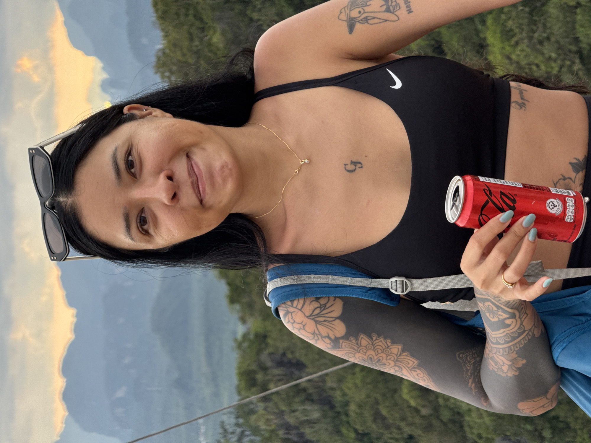 A woman with dark hair and tattoos on her arms and chest stands in front of a mountain range with a sunset sky. She is wearing a black sports bra and sunglasses on her head. She is holding a can of Coca-Cola in her right hand. The background shows lush green trees and misty mountains, characteristic of the landscape in Ban Khao Tang, Thailand. The lighting suggests it is late afternoon or early evening.