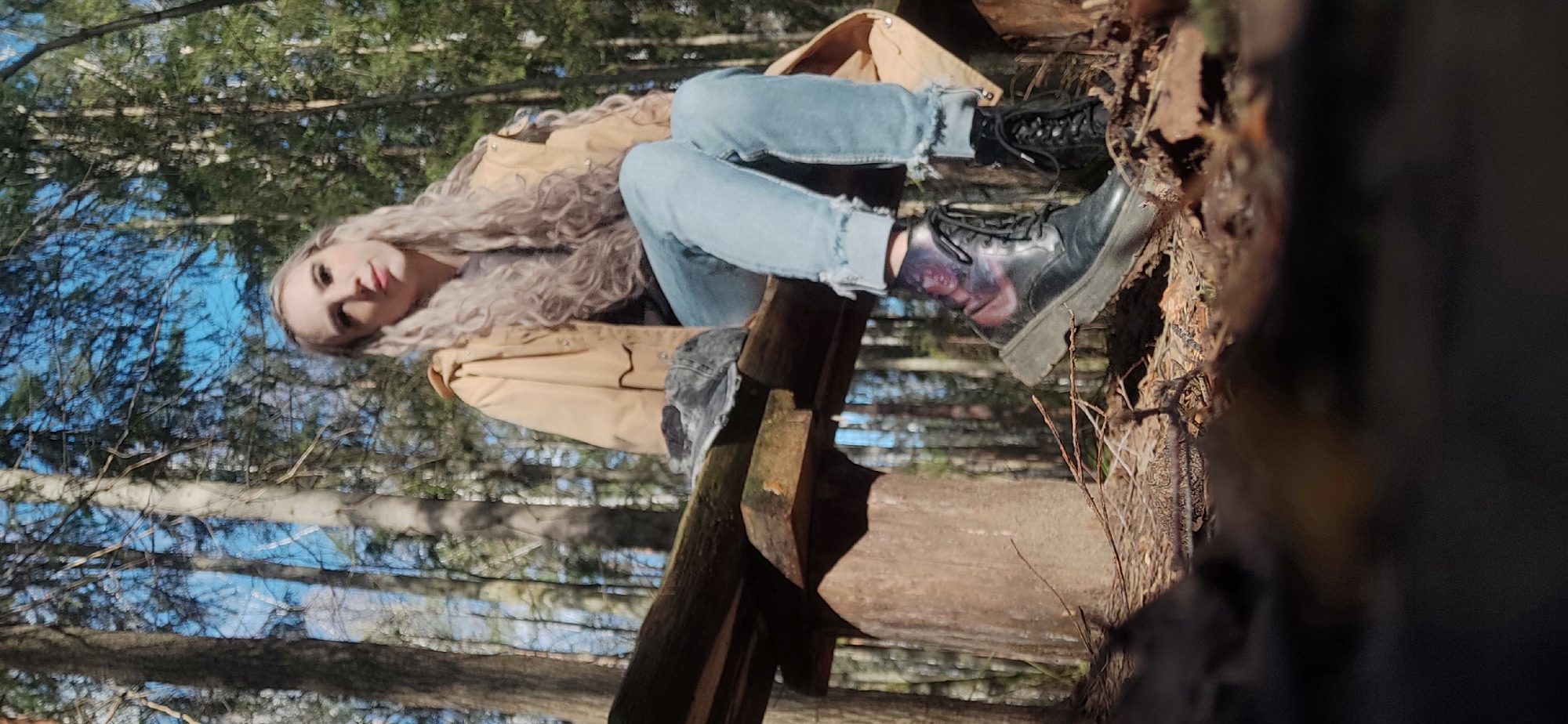 A person with long, light-colored, curly hair is sitting on a wooden structure, possibly a broken bench or a small bridge. They are wearing light blue jeans with frayed cuffs, a tan jacket, and black boots. One of the boots appears to have a design on it. The location seems to be a forest, with tall trees and blue sky visible in the background. The exact location and city are unknown.
