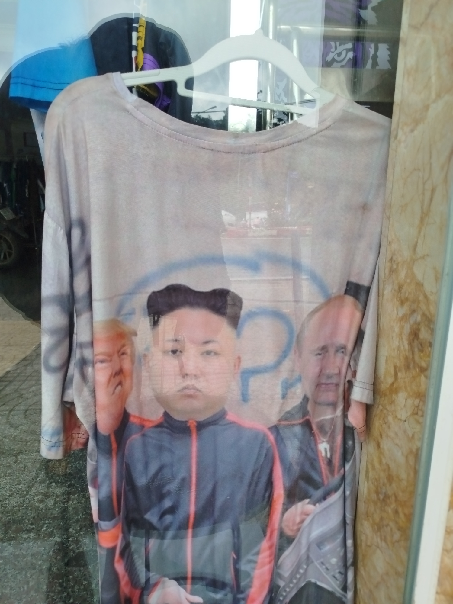 A t-shirt featuring a print of Kim Jong Un, Donald Trump, and Vladimir Putin is hanging on a white hanger in what appears to be a shop window. The t-shirt is light grey with an all-over print depicting the three political figures. Kim Jong Un is in the center, with Donald Trump to his left and Vladimir Putin to his right. They are all depicted wearing athletic jackets. The t-shirt is partially covered by a reflection of the surroundings, which includes what looks like a marble-patterned wall to the right and a blurry street scene or interior in the background. The image was taken in Da Lat, Vietnam, likely in a commercial area or market.