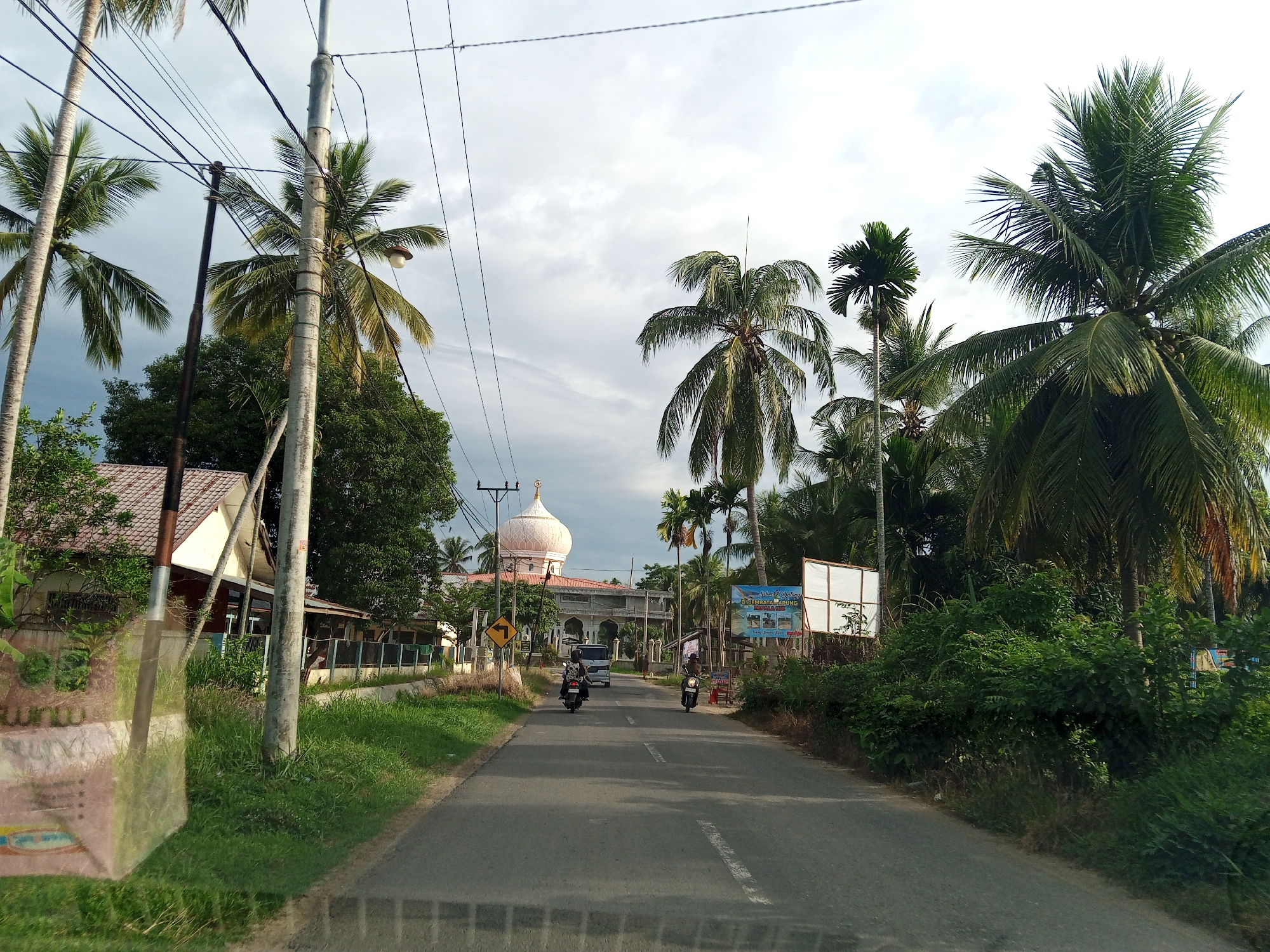 This image captures a view down a two-lane asphalt road in Pantebaroglesiblah, Indonesia, leading towards a mosque with a light pink dome in the distance. The road displays a dashed white centerline. Two individuals operating motorbikes are observed on the road, traveling away from the viewer, with a white van further ahead also moving in the same direction.
The environment is characterized by abundant tropical vegetation. Numerous tall palm trees, including coconut palms and what appear to be areca palms, flank both sides of the road and extend into the background. Dense green bushes and smaller trees line the road shoulders.
On the left side of the road, a single-story building with a reddish-brown tiled roof is partially visible behind a cluster of utility poles. Multiple utility poles, some concrete and others dark metal, support numerous black overhead power lines stretching across the scene. A street light is affixed to one of the concrete poles. A yellow diamond-shaped traffic sign, indicating a sharp turn or curve, is also present on the left.
On the right side, a large, rectangular white billboard frame stands, partially obscuring a smaller sign that includes the text "Selamat Datang di Tembara Bahung." Beyond this, dense foliage and palm trees obscure further details.
The sky is overcast with a mix of grey and white clouds. The lighting indicates daytime conditions.