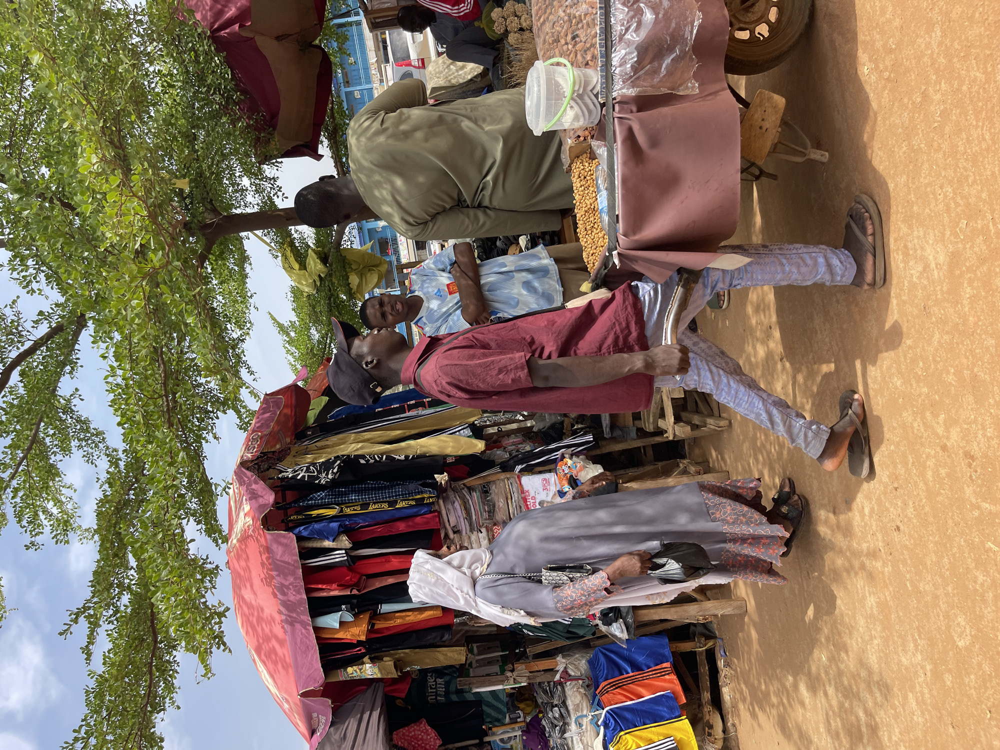 In Zaria, Nigeria, a street scene shows several individuals near market stalls. One stall displays stacked clothing under a red canopy. A woman in a gray dress and white headscarf is looking at the clothes. Nearby, a man in a red shirt and baseball cap walks past. At another stall, a man in an olive-green shirt stands with two other men. This stall has nuts and a clear container of items for sale. The ground is reddish. A tree with green leaves provides some shade to the area.