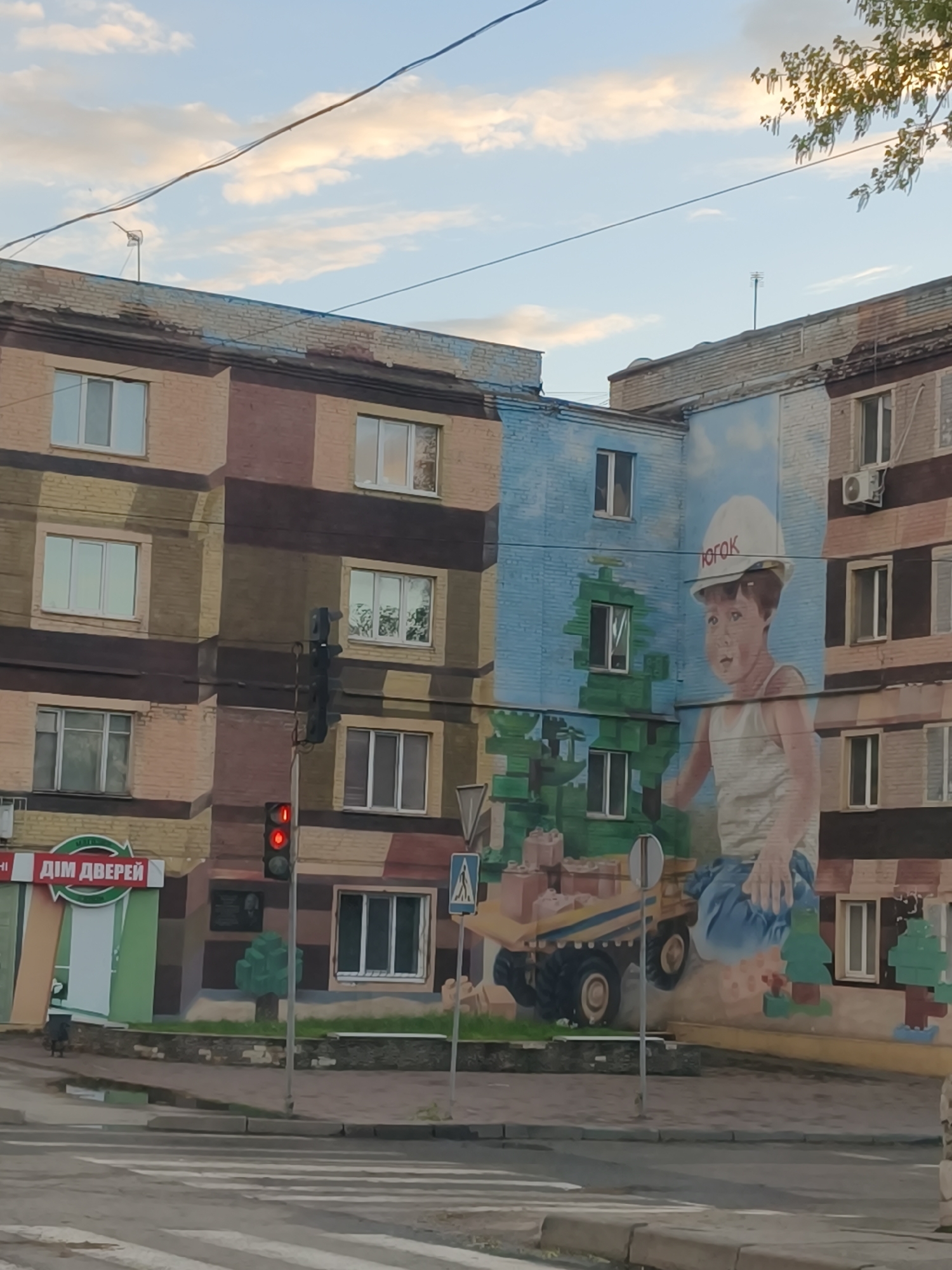 The image shows an intersection in Kryvyi Rih, Ukraine. On the corner of a multi-story building is a large mural depicting a young boy wearing a hard hat with the word "ЮГОК" (YUGOK) printed on it, sitting atop a toy dump truck constructed from building blocks. The building adjacent to the mural is painted in a camouflage pattern. A traffic light is visible showing a red light. A store sign "Дім Дверей" (House of Doors) is on the building. A pedestrian crossing sign stands near the mural. A portion of a tree with green leaves is visible in the upper right corner of the image, and power lines are strung across the sky.