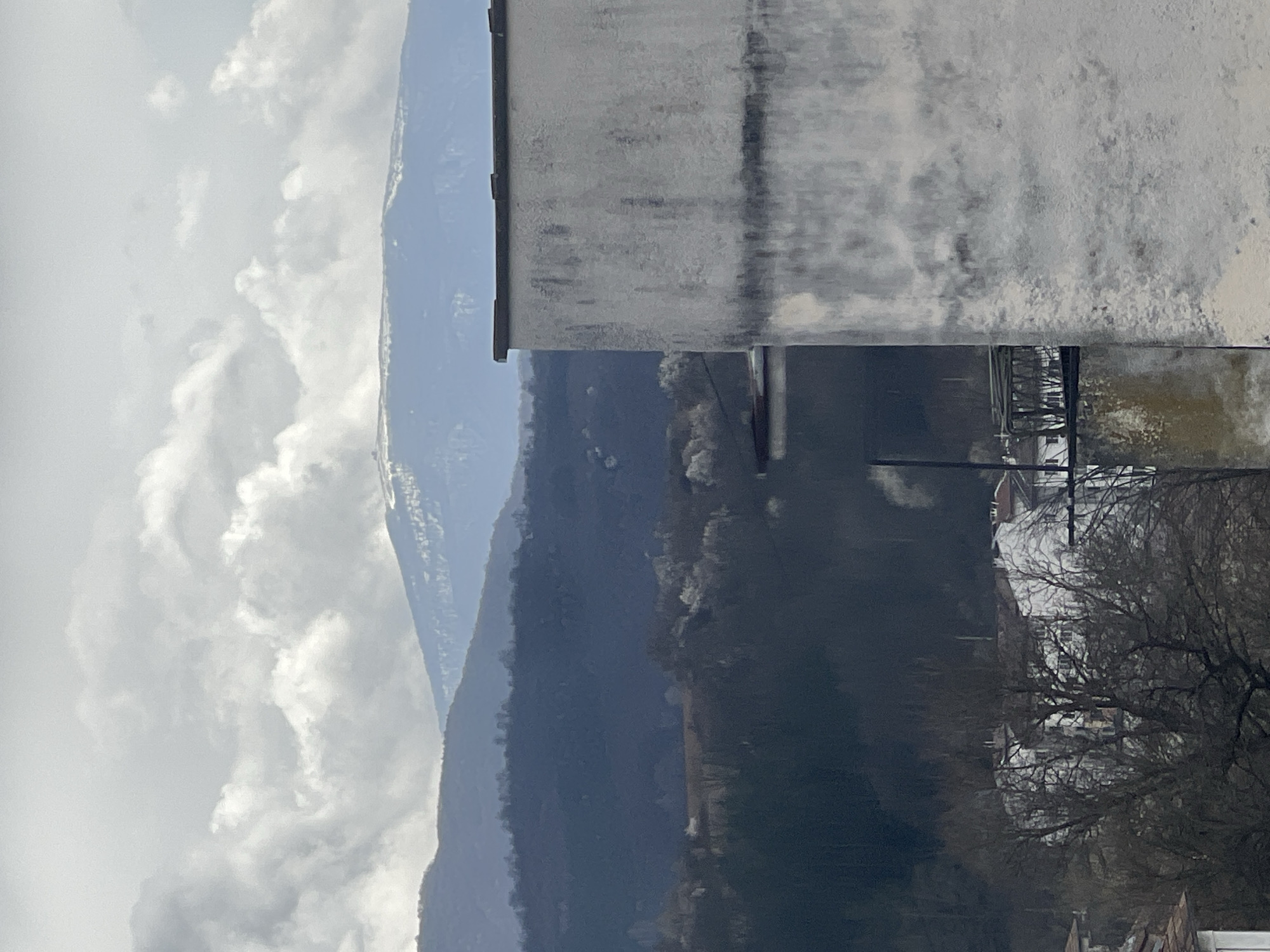 The image displays a panoramic landscape view over Elena, Bulgaria. The right foreground features a heavily weathered, light-grey building wall exhibiting dark vertical streaks and discoloration, topped by a dark, flat coping. Below and to the left of this, a dark, flat architectural element is partially visible. The midground reveals a valley floor and hillsides densely covered with primarily bare deciduous trees and some evergreens, interspersed with patches of lighter-colored vegetation, potentially snow-covered. Within this area, scattered white residential buildings with red roofs are discernible. The background consists of multiple successive ranges of mountains, with the farthest peaks exhibiting visible snow cover. The sky is filled with a mix of white and grey cumulus clouds, indicating an overcast or partly cloudy day. No human subjects or active events are present.