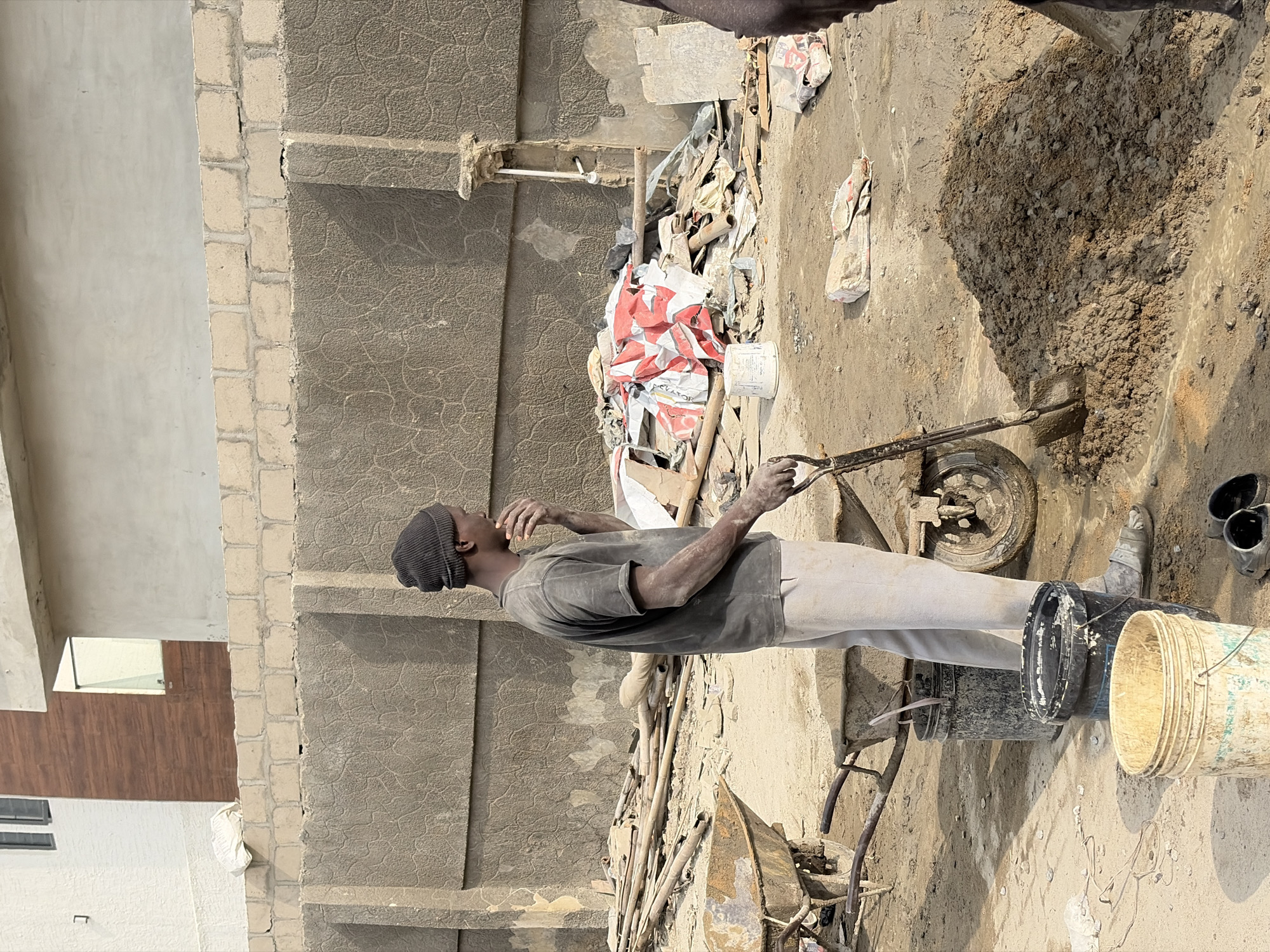 A man wearing a dark beanie and a dusty grey t-shirt stands in a construction site in Igboefon, Nigeria. He is in the process of mixing cement with a shovel, his hand resting on his chin as he looks to the right. The ground is covered in dirt and debris, with two wheelbarrows and several buckets scattered around. In the background, a textured concrete wall separates the construction site from a building under construction. The overall scene depicts a typical day of manual labor in a developing urban environment.