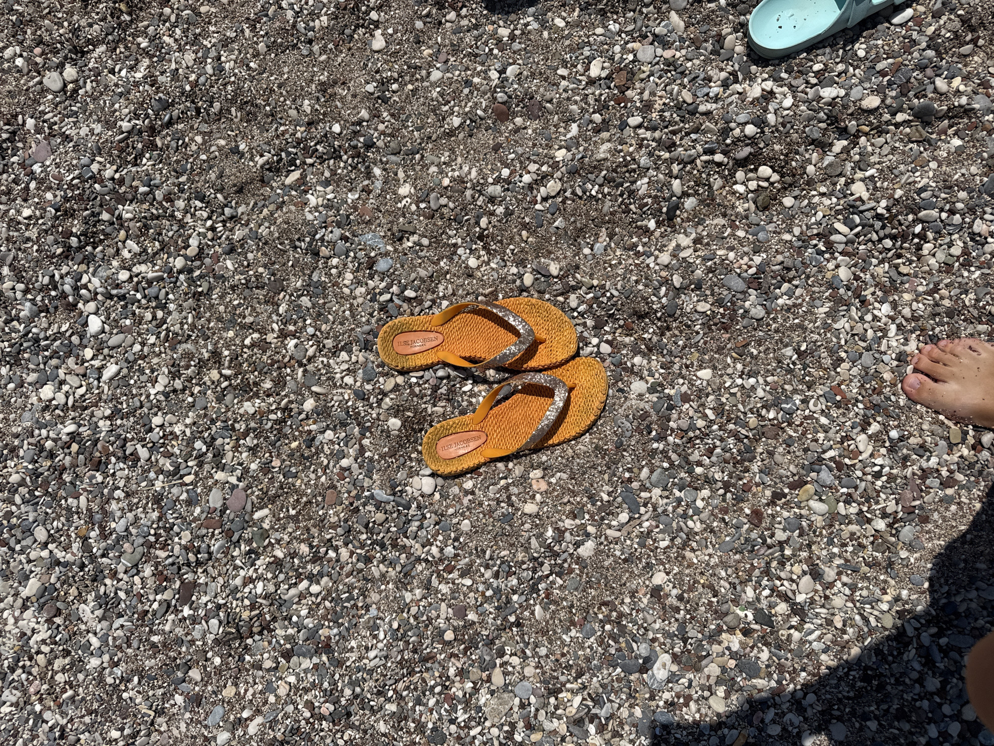 A pair of orange flip-flops with glittery straps sits on a pebbly beach. To the right, a portion of a light blue sandal is visible. In the lower left corner, a person's bare foot is visible, with a shadow cast over the sand and pebbles. The scene is set on a beach with an abundance of small, multi-colored pebbles.