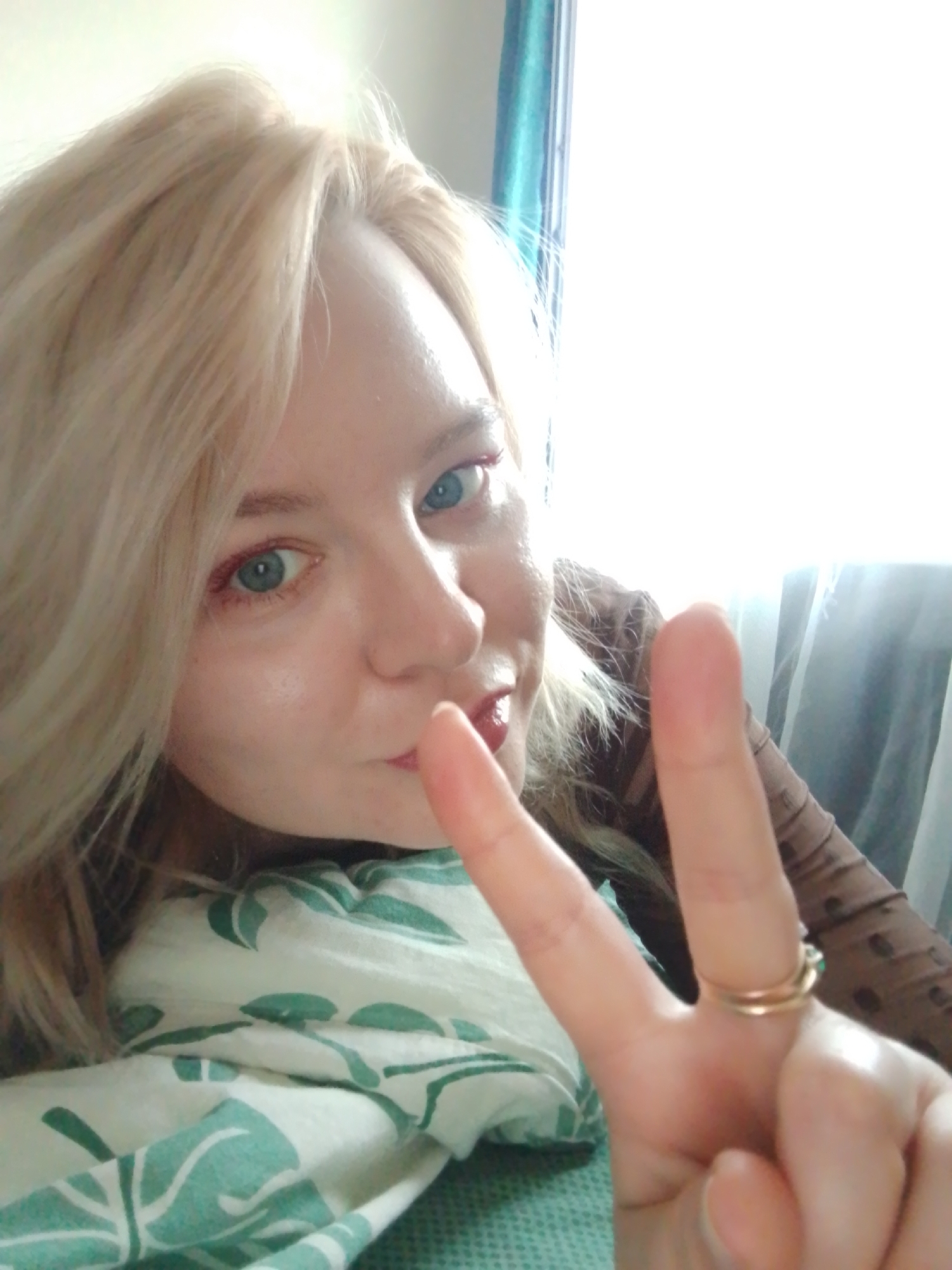 A blonde-haired woman with blue eyes is making a peace sign, she has on red eye shadow and a gold ring. The picture is taken indoors and the woman is lying in bed with green and white patterned sheets. The background is a window with white curtains. The image does not provide any information to indicate the location of Izhevsk, Russia.