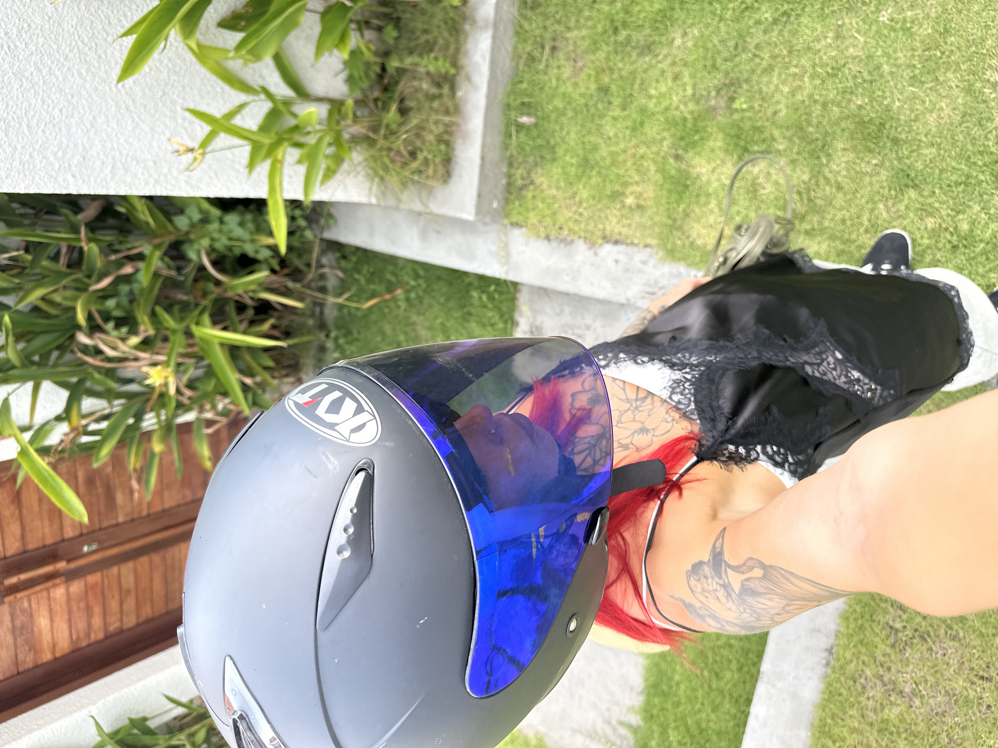 A woman with red hair and multiple tattoos is seen from an overhead perspective wearing a black and white dress and black shoes. She is holding a gray motorcycle helmet with a blue visor. The background consists of green grass, plants, and part of a wooden structure. The image was taken in Dalung, Indonesia.