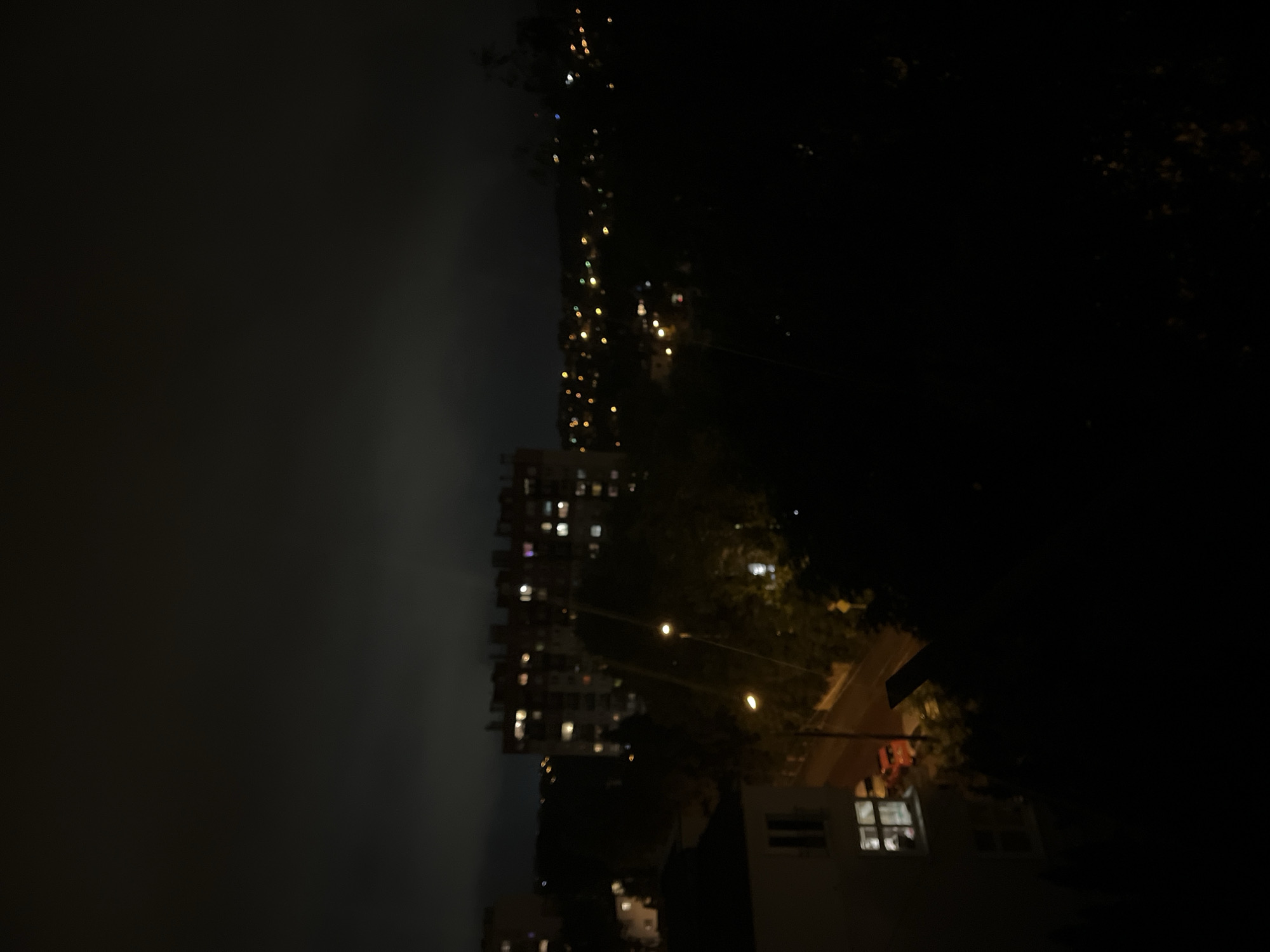 The media file displays a nighttime scene, appearing to be an urban or suburban landscape. The original image is oriented vertically, but the content suggests a horizontal perspective when rotated 90 degrees counter-clockwise.
In this re-oriented view, the upper portion of the image reveals a dark sky, which appears hazy or misty, possibly indicating cloud cover or atmospheric conditions. Below the sky, on the left side, stands a tall, multi-story building, likely an apartment complex, with numerous windows illuminated, suggesting active residences within. The structure of this building is visible but indistinct due to the low light.
To the right of the tall building, and extending across the middle and right side of the frame, are dark silhouettes of trees. Several artificial lights, likely streetlights, are visible among the foliage and in the distance. These lights appear somewhat blurred, creating a bokeh effect. In the lower right corner, a section of a smaller building is visible, featuring at least one brightly lit window.
No people are discernible, and no specific activities are taking place. The overall impression is one of a quiet night in a populated area. The image quality is somewhat soft and grainy, consistent with low-light photography. No visible text is present.