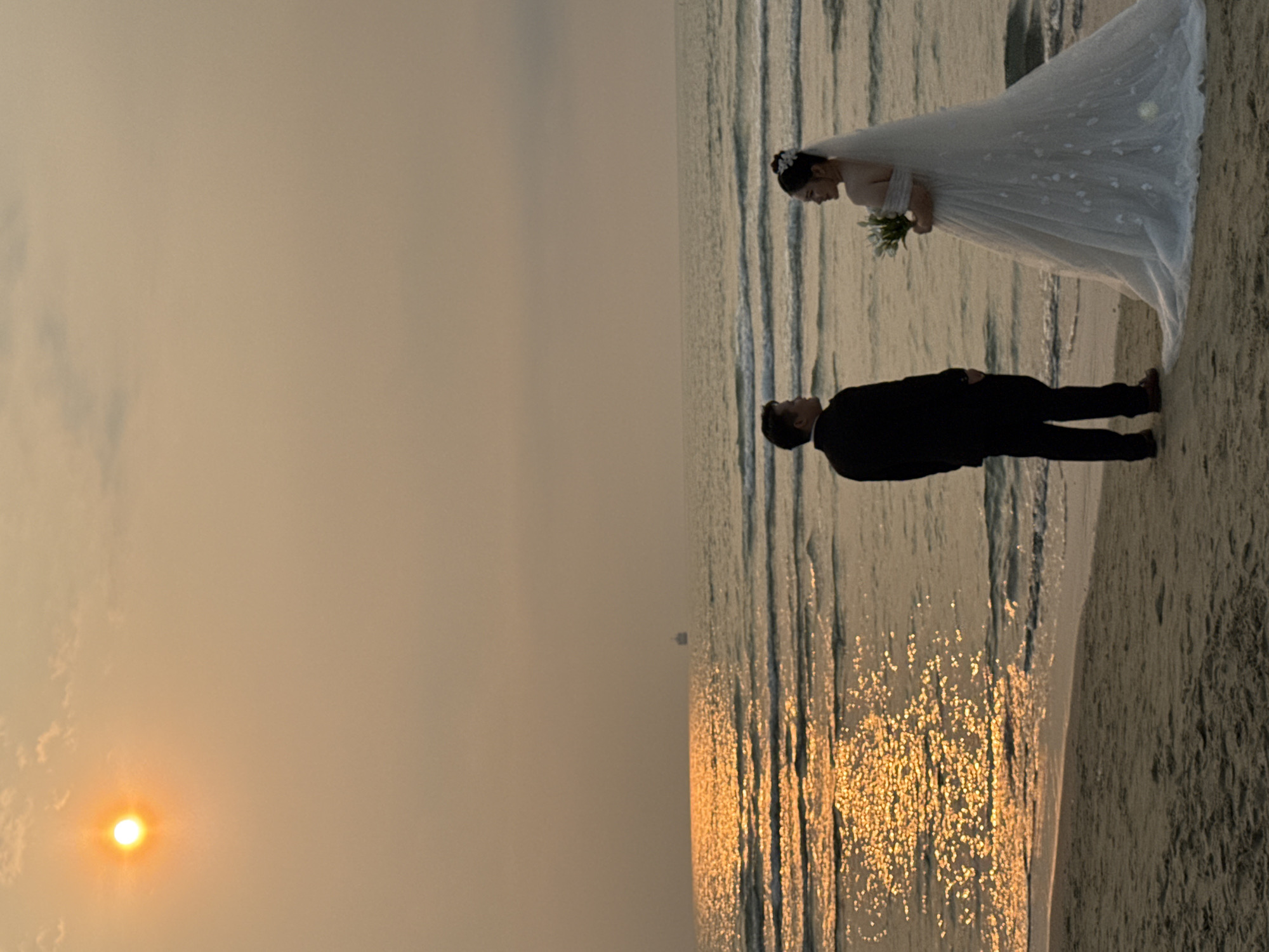 A photograph captures a wedding couple on a beach in Đà Nẵng, Vietnam, at sunset. The bride, wearing a white wedding gown and veil, holds a bouquet. The groom is dressed in a black suit. They are standing on the sand, with the ocean stretching out behind them. The sun is setting on the left side of the frame, casting a golden light across the water and sky.