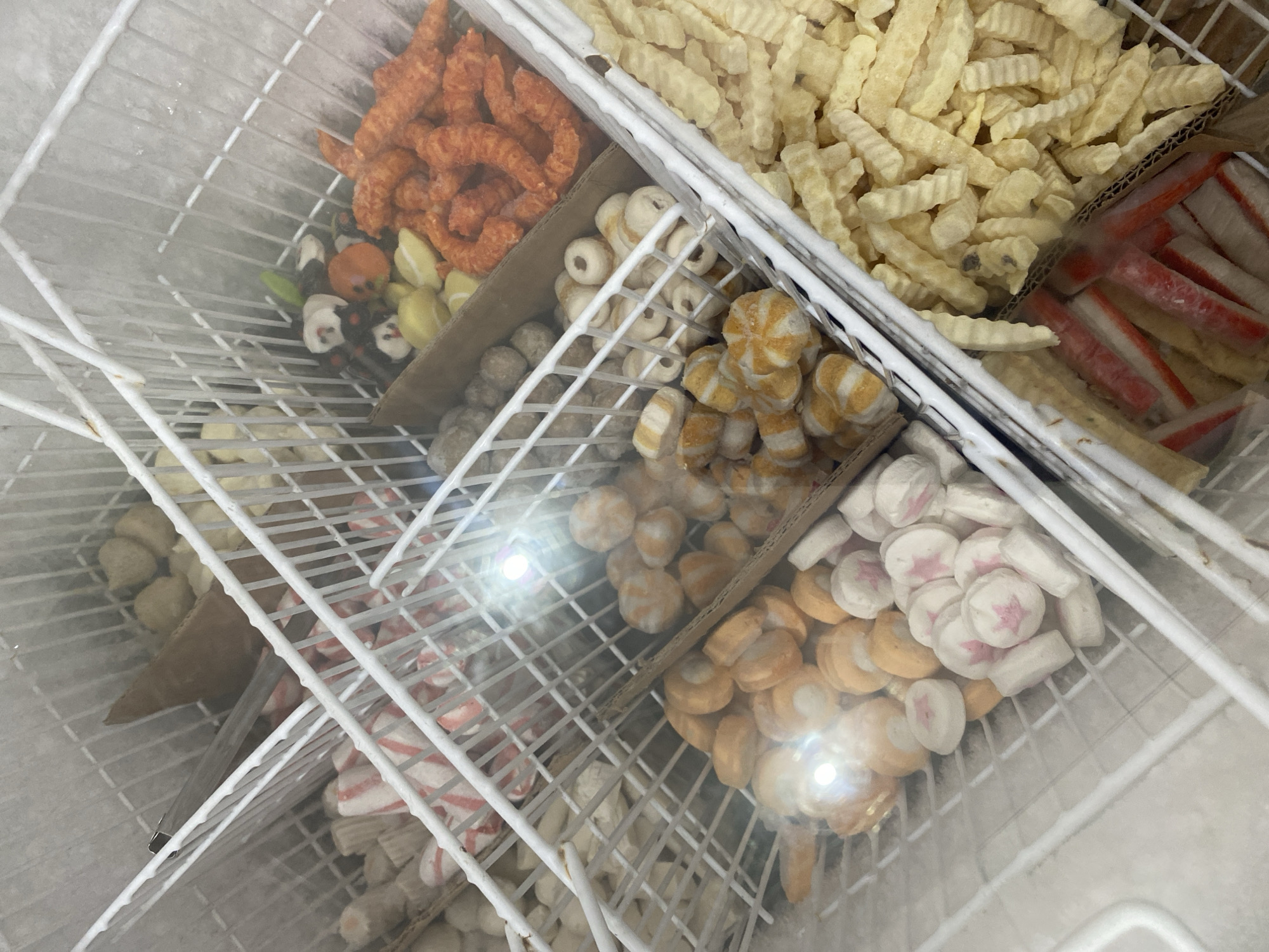 The image shows a frozen food display in a store, likely a supermarket or a specialized frozen food shop, located in Payatieng, Indonesia. The display is organized into white wire baskets that divide the frozen items into different compartments.
The contents visible include:
- Various types of frozen seafood products, such as fish balls and imitation crab sticks.
- Some brightly colored orange, curled, processed food items.
- Crinkle-cut french fries.
- What appears to be a piece of sweet potato or yam.
- Other unidentifiable frozen goods.
The environment is cold, as evidenced by the frost on the interior of the display unit. The lighting is artificial and there are reflections on the transparent cover of the display, indicating a commercial setting. There are no people visible in the image. The overall impression is of a well-stocked frozen food section.