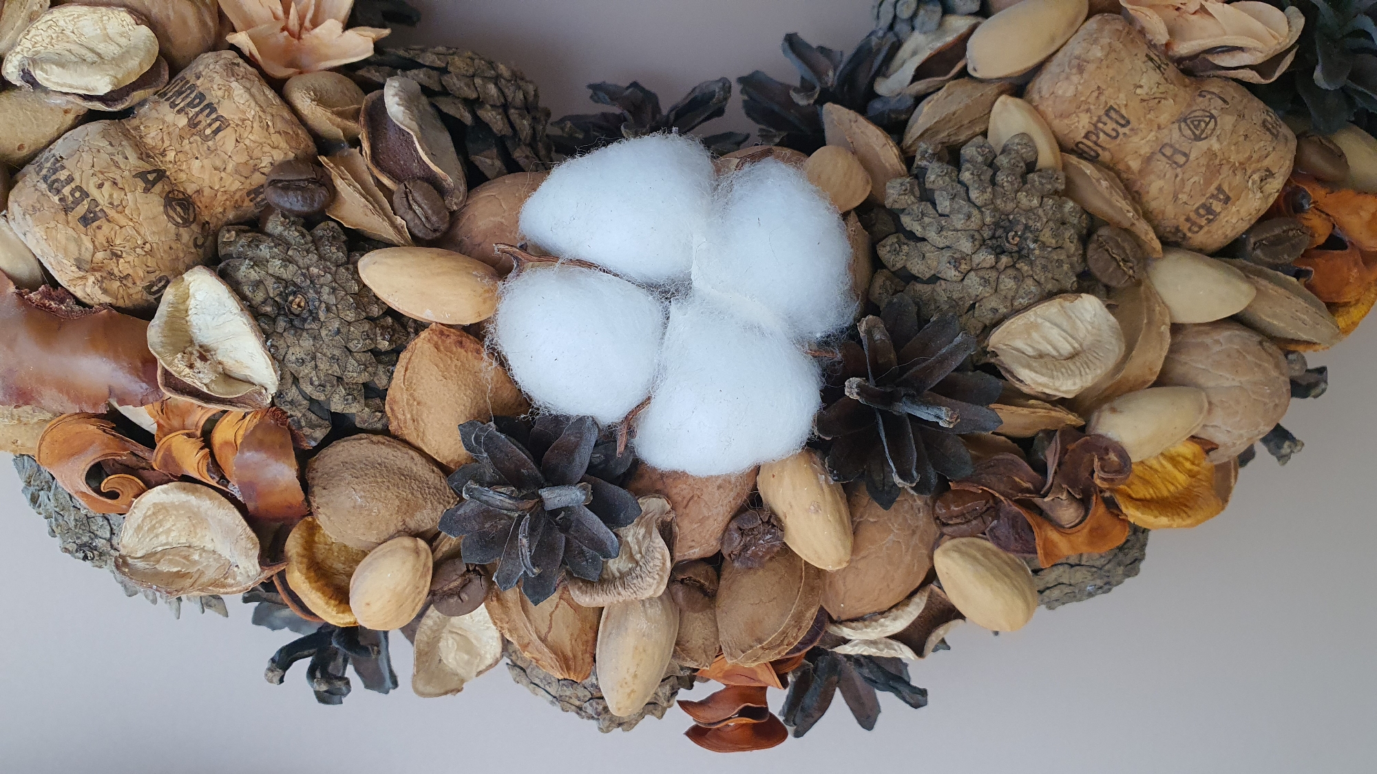 A close-up, overhead shot of a decorative wreath is presented against a plain, light-colored background. The wreath is composed of an assortment of natural elements, including nuts, dried seed pods, pine cones, dried flowers, and coffee beans, all arranged in a circular fashion. A prominent cluster of white cotton is visible near the center of the frame. The items are densely packed, with a variety of textures and earthy tones dominating the color palette. The cork of a wine bottle, stamped with Cyrillic text, is also incorporated into the wreath. The image does not provide any information about the city of Krasnoyarsk, Russia.