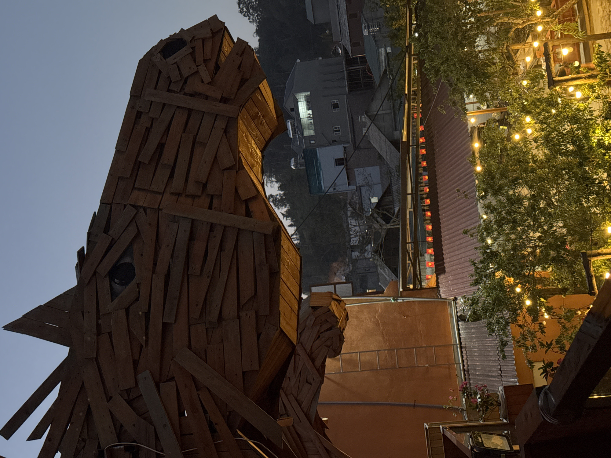 The image, taken in Bắc Hà, Vietnam, features a large wooden sculpture of a rooster in the foreground. The sculpture is constructed from rough wooden planks and appears to be a prominent landmark.
In the background, buildings and structures characteristic of the city are visible. A building constructed from lighter-colored concrete and painted a lighter shade is seen in the distance. There are also buildings with red roofing. Decorations with lights and flags can be seen around the building.