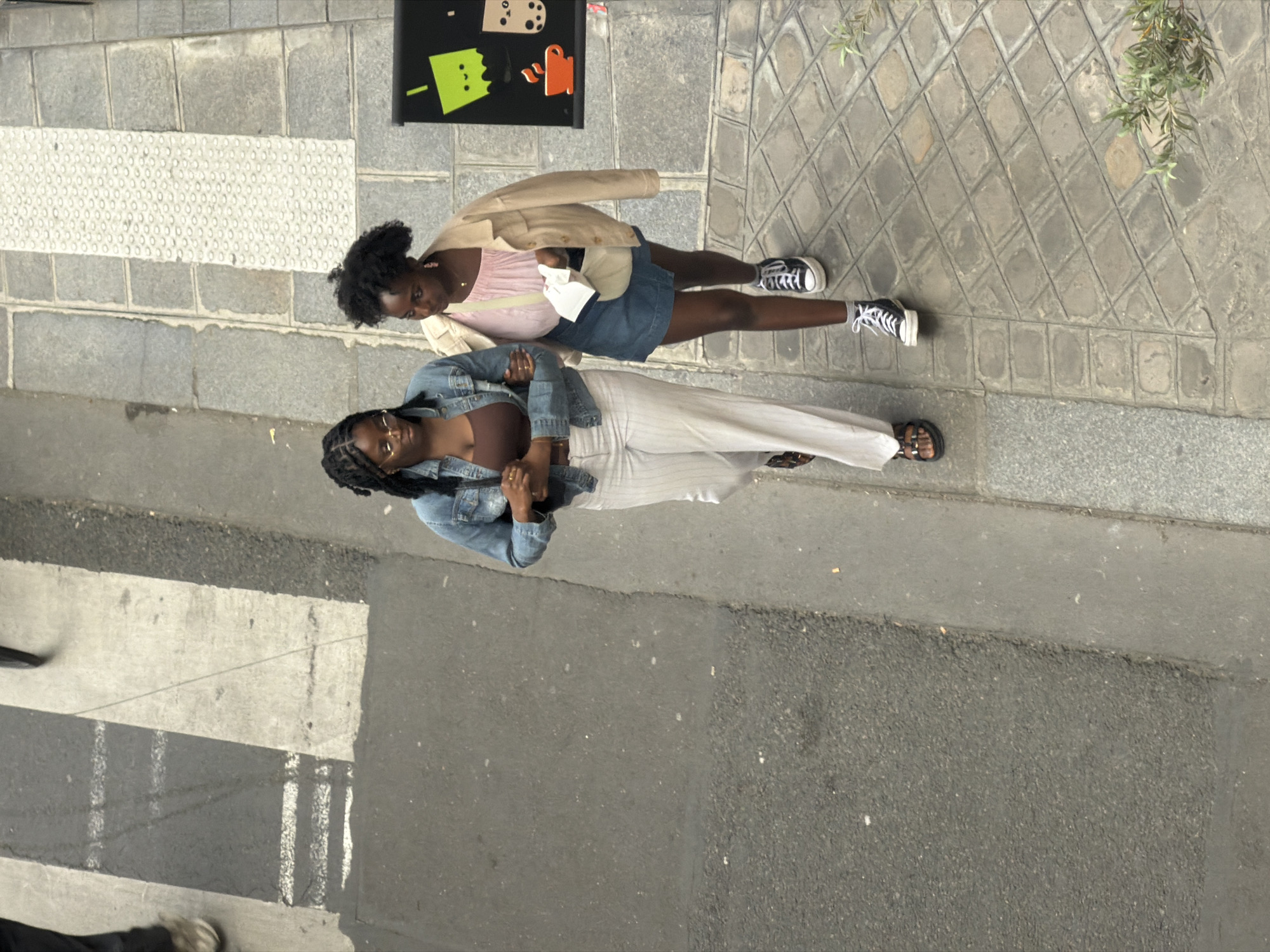 The image shows two young women standing on a city street, likely in Paris, France. The women are photographed from a high angle, looking down. One woman is wearing a light pink top, a denim skirt, a beige jacket, and black Converse sneakers. The other woman is wearing a brown top, white pants, a denim jacket, glasses, and brown sandals. They are standing on a sidewalk paved with stones and asphalt. There is a street sign for a bubble tea store. In the background, there are trees and foliage. The street has zebra crossing markings. The lighting suggests it is daytime. The environment is urban and appears to be a pedestrian area.