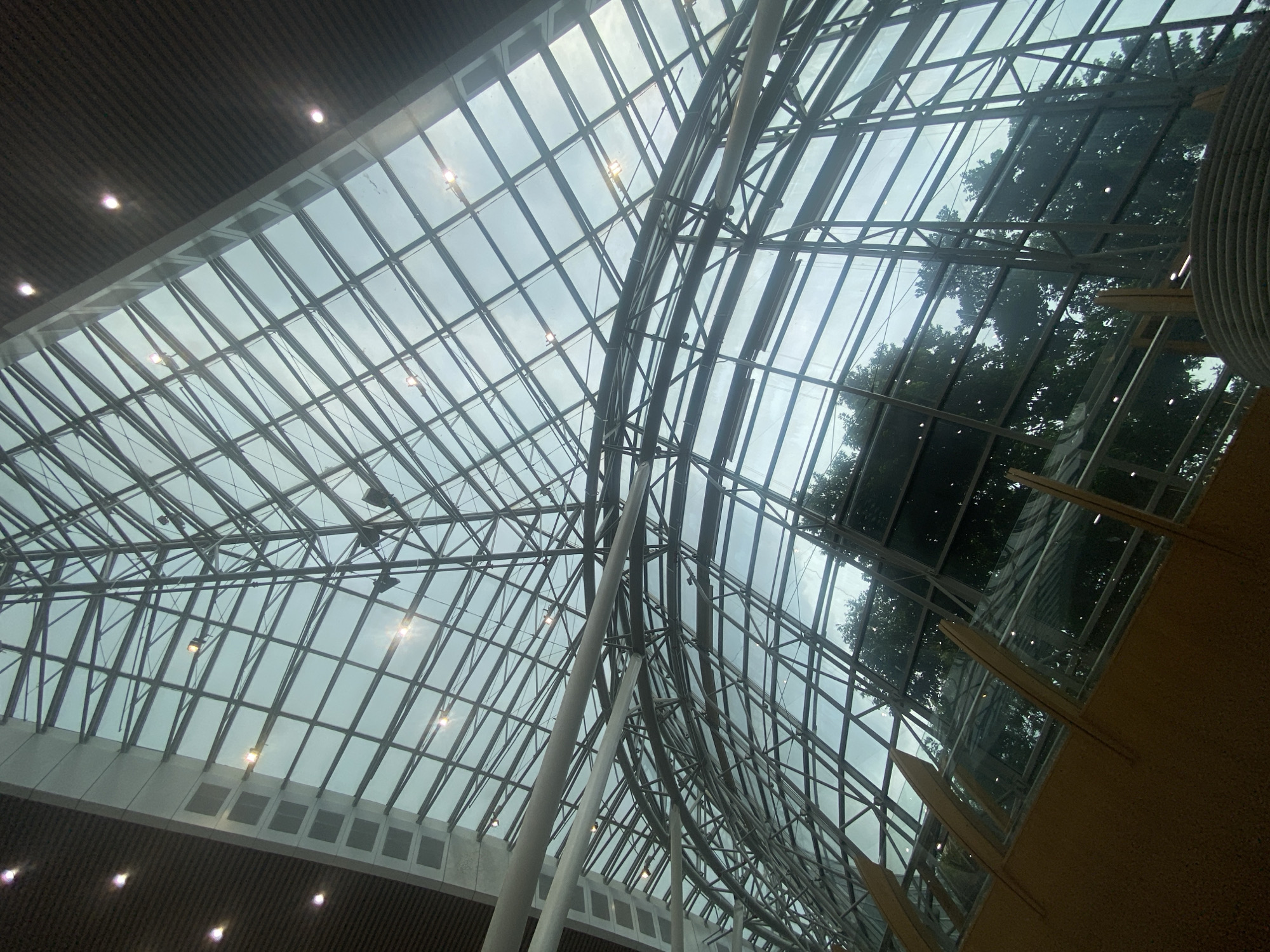 The image shows the interior of a modern building with a large, arched glass ceiling supported by a metal framework. The ceiling is a complex geometric pattern of intersecting beams and panels, allowing natural light to filter through. Trees are visible through the glass, suggesting the building is surrounded by greenery. The lower portion of the image shows a wooden structure with angled supports, possibly part of the building's interior architecture or seating. The lighting appears to be a mix of natural light from the sky and artificial lights embedded in the ceiling. The overall impression is one of spaciousness and contemporary design. The location is identified as the KLIA Aerotrain station in Sepang, Malaysia.
