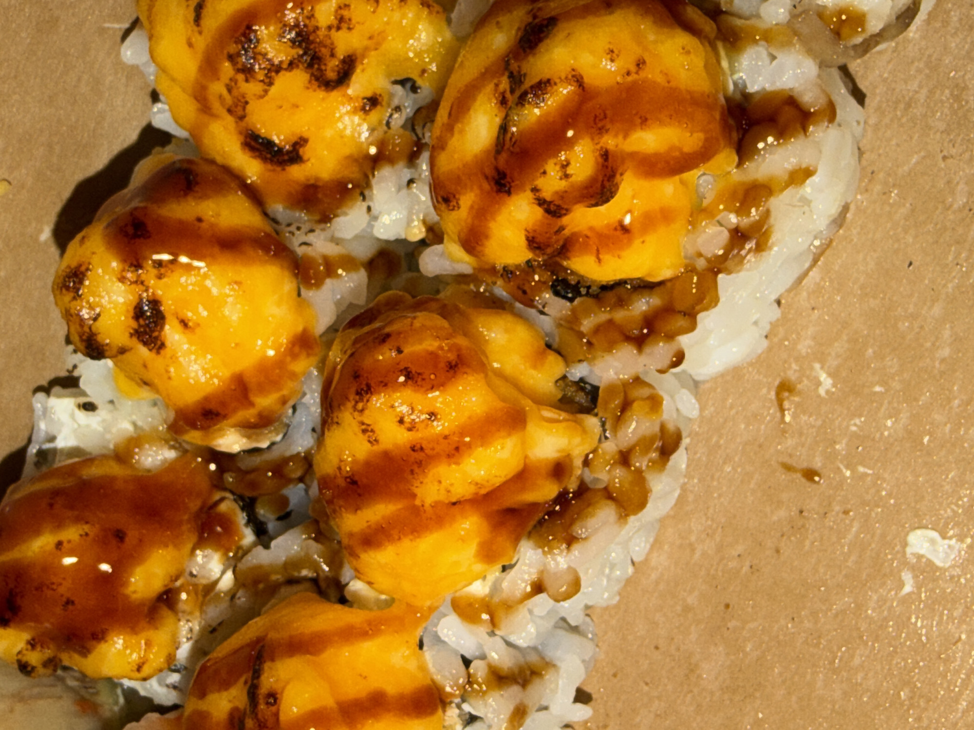 The image shows a close-up view of a serving of sushi rolls, likely in a takeout container. The sushi appears to be topped with a yellow-orange sauce that has brown lines and spots, possibly from grilling or searing. The rolls are placed on a brown surface. The city of Varna, Bulgaria is not identifiable from the image.