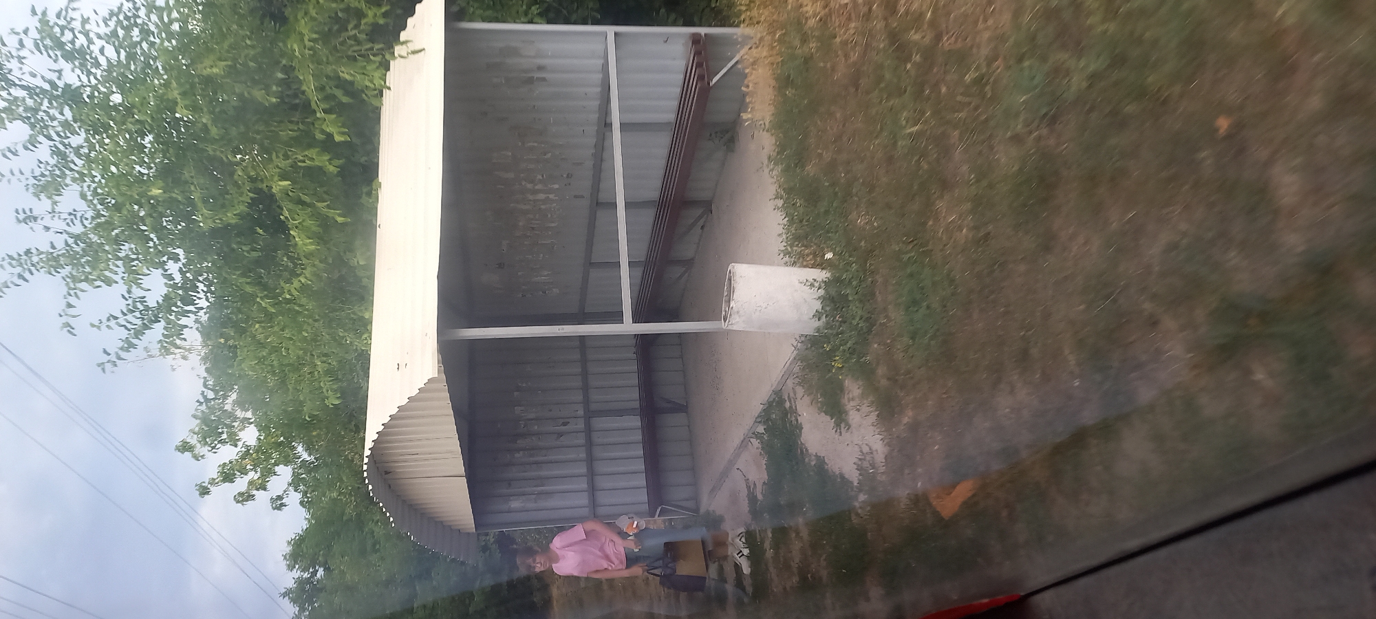 A woman in a pink shirt and blue jeans stands near a bus stop shelter, holding a large brown bag. The bus stop has a corrugated metal roof and sides, with two wooden benches inside. A cylindrical concrete trash can sits next to the shelter. The ground is covered with dirt and patchy grass. Trees and foliage surround the bus stop. The scene appears to be located in a rural or semi-rural area on the outskirts of Klyntsi, Ukraine. The image is taken from inside a vehicle, as evidenced by the reflection on the lower left of the frame.