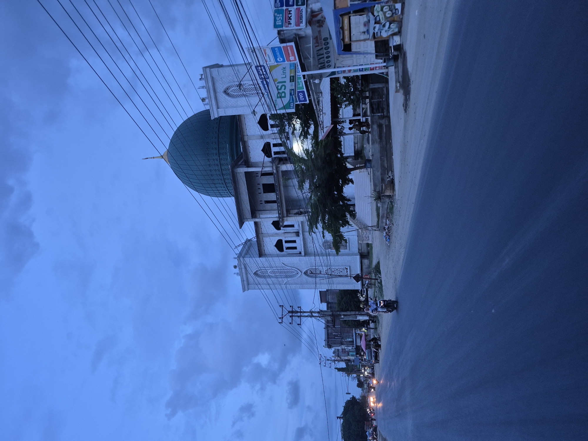 The image shows a street scene in Keudelapang, Indonesia, taken during twilight or dusk. A white multi-story building with a large, turquoise dome stands prominently on the left. Signboards for "BSI Link" and other businesses are visible on the buildings to the right of the mosque. Electrical wires crisscross the sky above. A paved road stretches into the distance with faint reflections from streetlights and vehicle headlights. Several motorcycles are on the road. The sky is blue with scattered clouds. A tree is growing in front of the mosque.