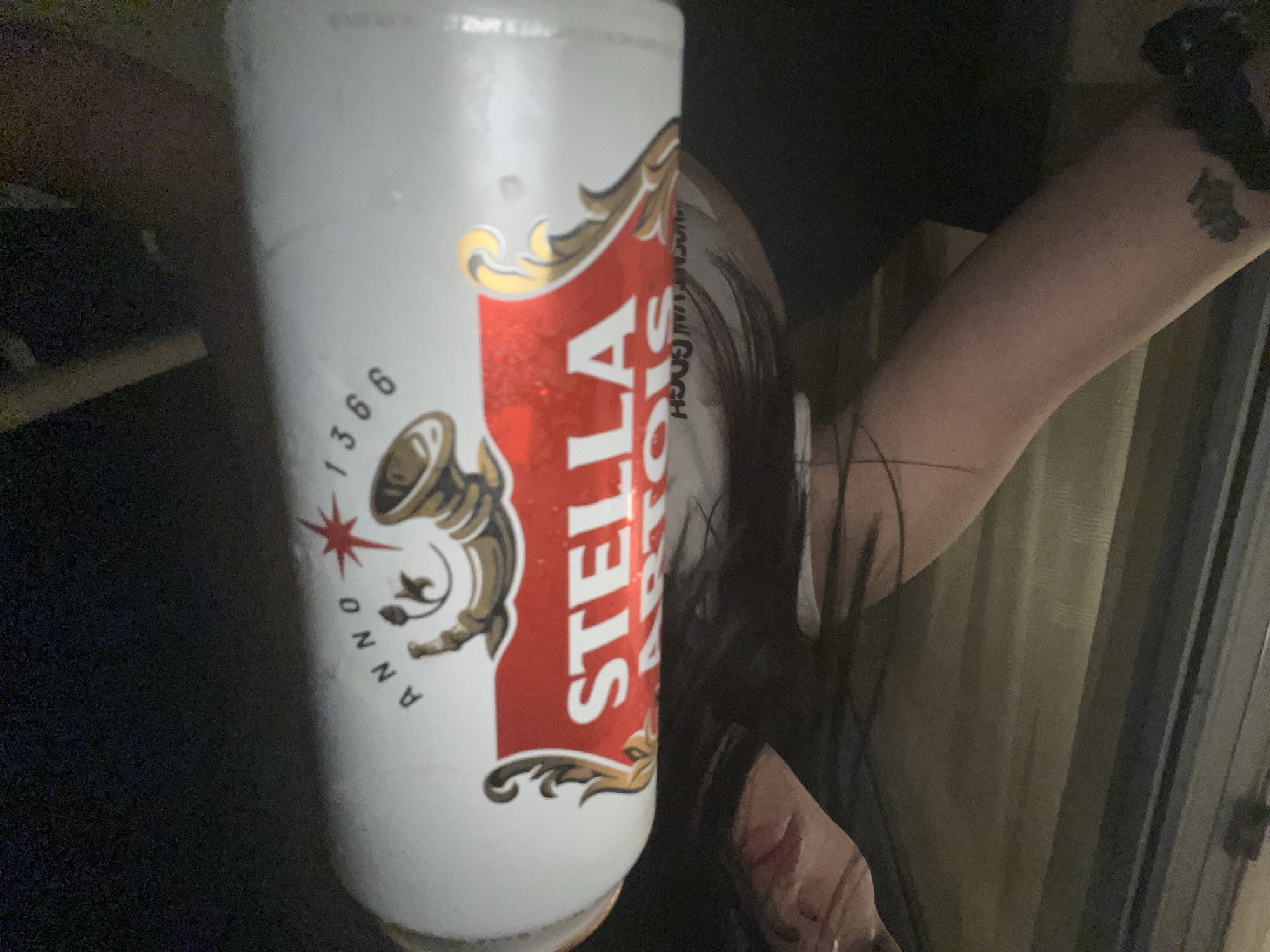 The image, rotated 90 degrees counter-clockwise, presents a close-up view of a Stella Artois beer can held by a person in a dimly lit indoor environment. The white can prominently features the red and gold Stella Artois logo, which includes a star and a horn emblem. Below the emblem, the text "ANNO 1366" is visible. Notably, above the main "STELLA ARTOIS" text, the black lettering "VAN GOGH" is printed on the can, suggesting a special edition or collaboration. Condensation is visible on the can, indicating it is cold.
Part of a person's arm, hand, and dark hair are visible to the right of the can. A tattoo can be seen on the upper part of the arm, though its design is not clearly discernible. The background is dark and indistinct, with vertical lines on the right side that suggest curtains or window blinds. The setting appears to be casual, likely during the evening or night due to the low light conditions.