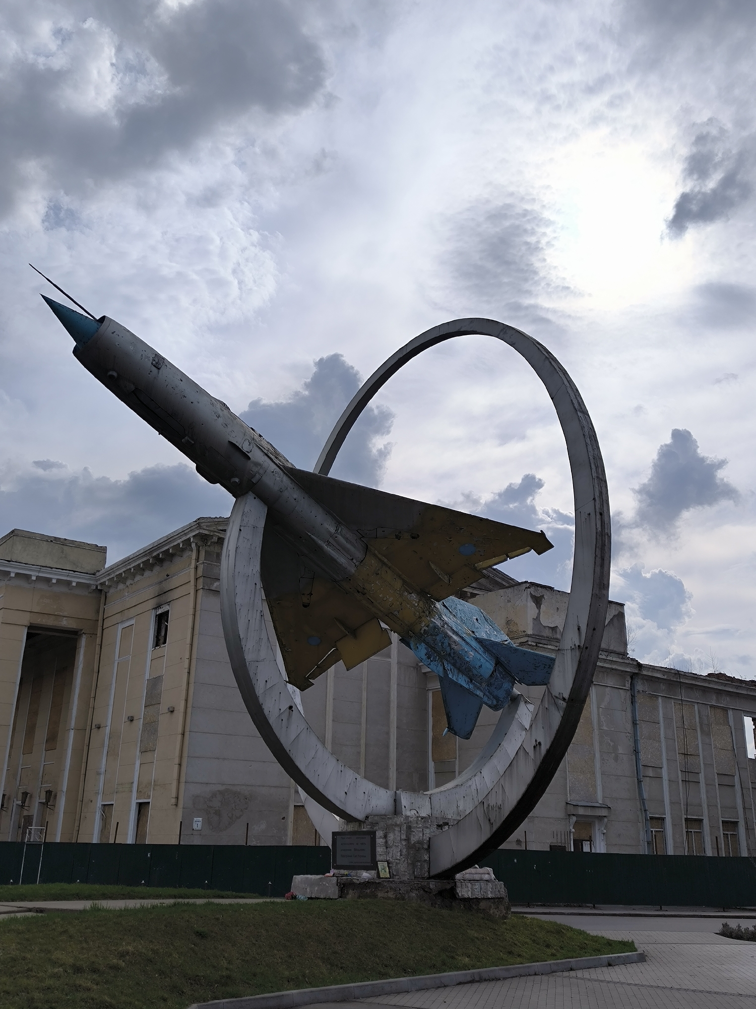 A low-angle, medium shot presents a MiG-21 fighter jet monument located in Vinnytsia, Ukraine. The aircraft, painted in muted grey, yellow, and blue tones, is mounted within a circular frame that bisects its fuselage. The nose of the jet, tipped with a blue antenna, points towards the upper left of the frame. The jet's tail section, painted primarily in blue, is visible on the right side of the frame.
The monument sits atop a small, grassy mound adjacent to a paved area. Behind the monument, a tall, derelict building stands, featuring classical architectural elements such as columns and a symmetrical facade. The building's facade is partially boarded up with some missing panes. A green barrier fence runs along the base of the building.
The sky is overcast, with a mix of dark and light grey clouds, obscuring the sun. The overall lighting is subdued, adding to the somber atmosphere of the scene.