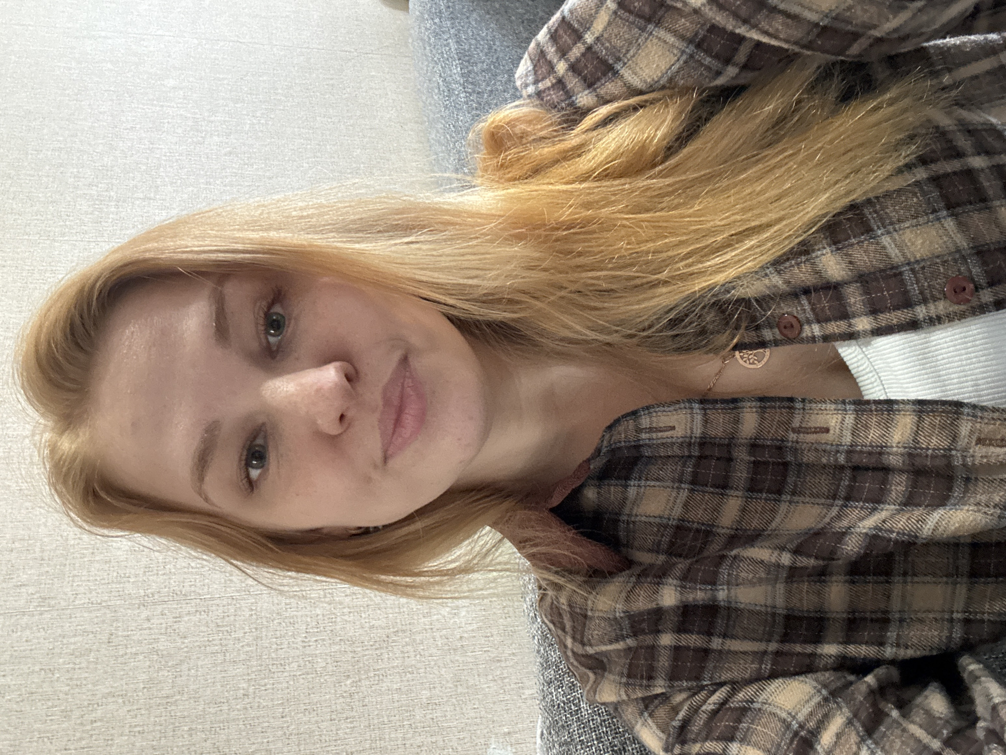 An indoor, eye-level shot, likely a selfie, captures a young adult woman with long, light-colored (blonde or strawberry blonde) hair parted to the side. She has fair skin, green eyes, and a neutral expression with a slight, gentle smile. The woman is wearing a brown and beige plaid flannel shirt, worn open over a white ribbed top. A gold-colored necklace with a tree-of-life pendant is visible around her neck.
Her hair flows over her shoulders and rests on her chest. The background features a light, textured wall on the left side and a grey upholstered couch or chair on the right. The lighting appears bright and even, suggesting an indoor setting during the daytime. The image is rotated 90 degrees counter-clockwise from a standard upright portrait orientation. No visible text is present in the image. The scene conveys a relaxed, casual atmosphere in what appears to be a home environment in Rusanovo, Russia.
