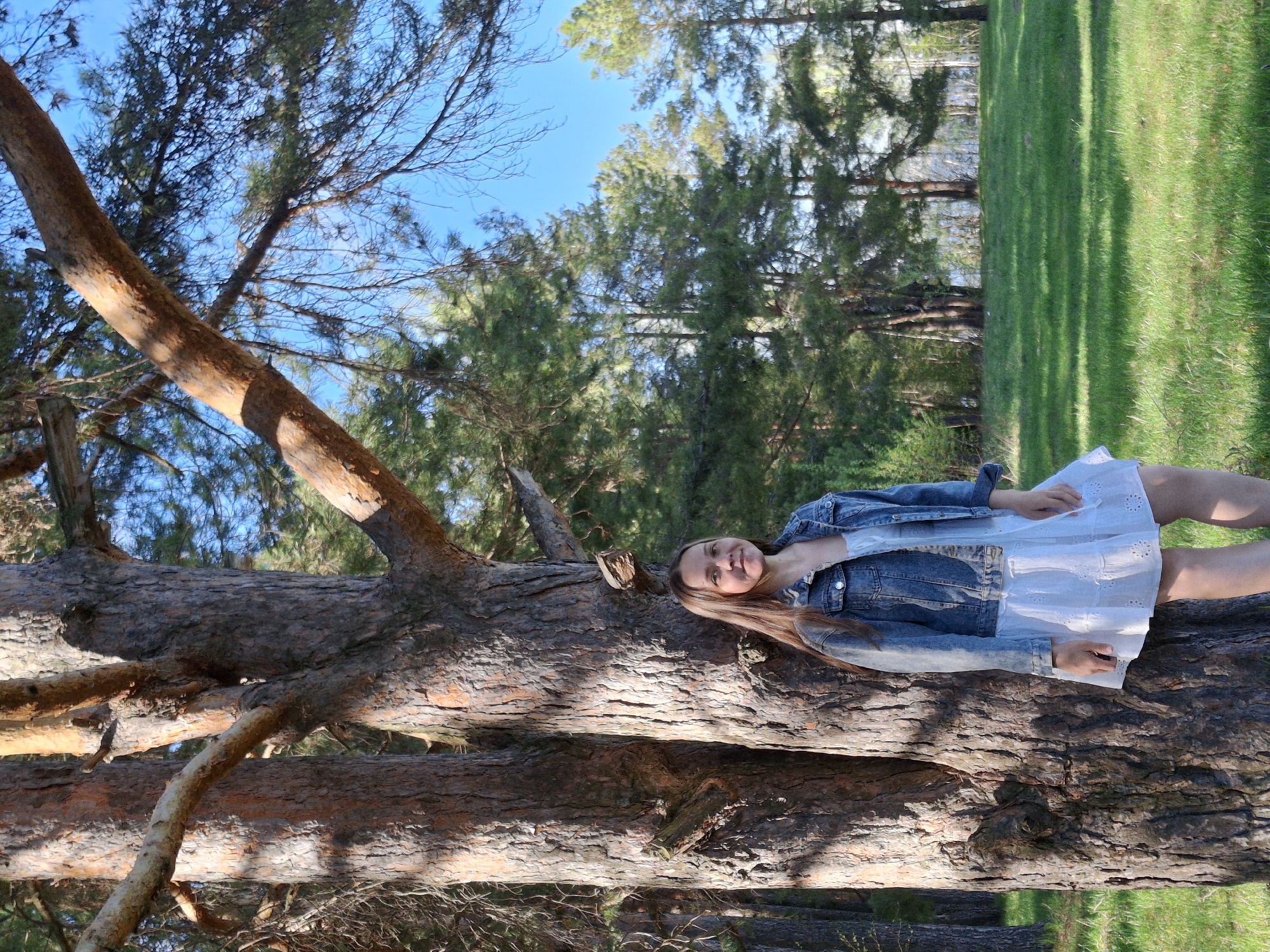 A young woman lies on a fallen tree trunk in a park-like setting. She is wearing a white dress and a denim jacket. Her long, brown hair is spread out on the tree trunk. She is smiling. The tree trunk is large and rough-textured, with visible branches. In the background, there are green trees, blue sky, and a grassy lawn. The image was taken outdoors during the daytime in Chaikovsky, Russia.