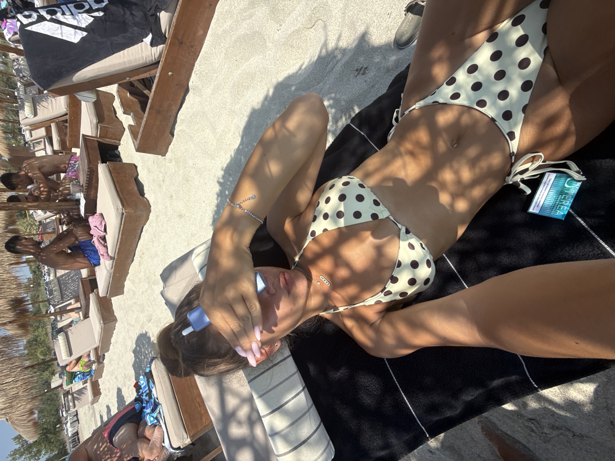 A woman lies on a beach lounge chair covered by a black towel, wearing a polka-dot bikini. She is shielding her eyes with her hand, which is holding a small blue object. Her nails are painted a pale pink. Sunlight casts dappled shadows across her body and the surrounding sand. Other beach lounge chairs, some with umbrellas, are visible in the background, along with other people relaxing on the beach. The scene suggests a sunny day at a beach resort. A pack of what appears to be mints with the brand name "Terea" is visible next to the woman. The location is identified as Katerini Municipality, Greece.
