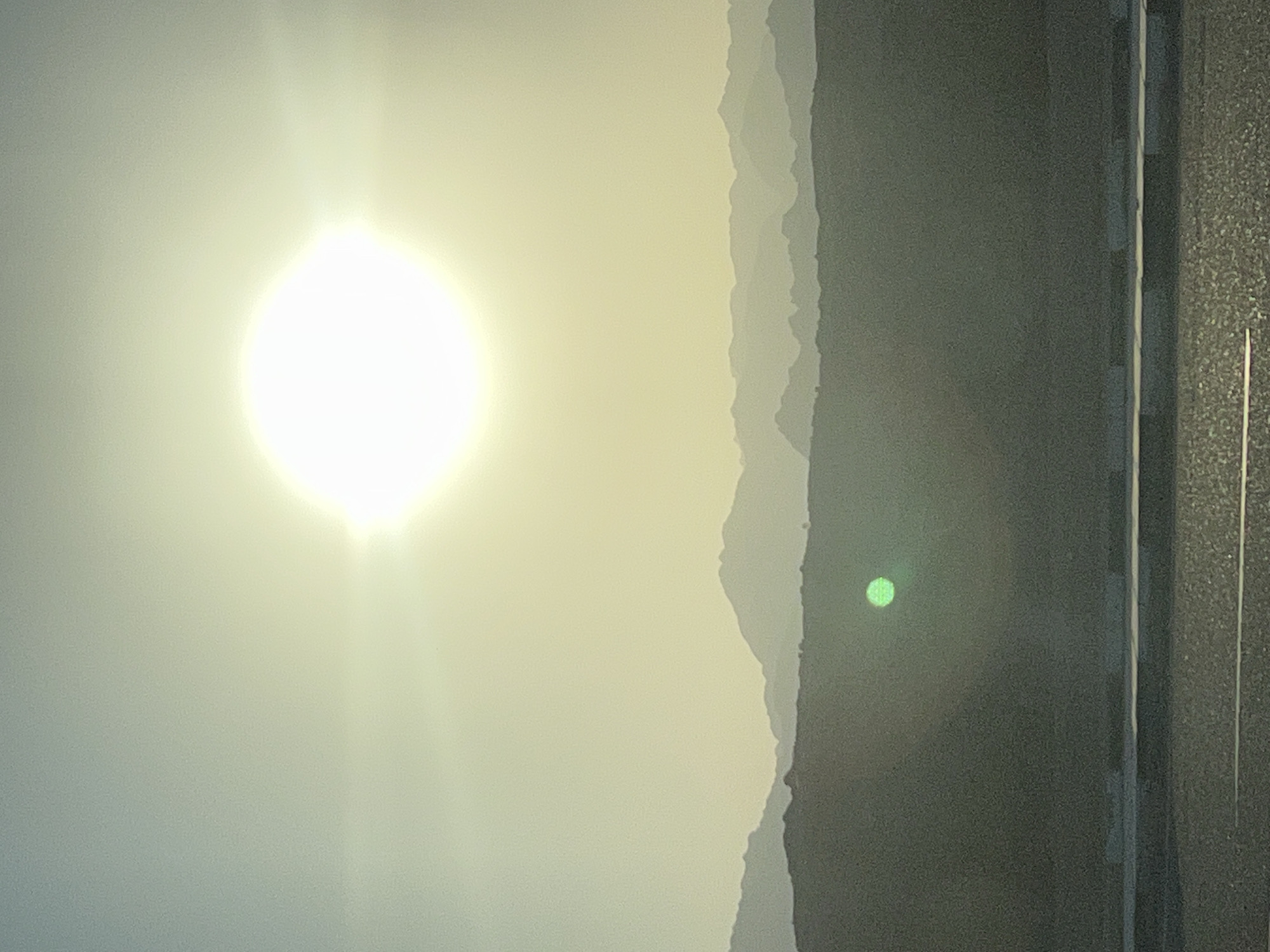 The image captures a desert landscape at sunset, likely near Sharm El Sheikh, Egypt. The sun, a large white orb with a bright halo, dominates the upper portion of the frame, casting a warm, hazy glow over the scene. In the middle ground, silhouetted mountain ranges stretch horizontally across the image, their forms softened by the atmospheric light. In the foreground, a dark, textured surface, possibly a road or pavement, stretches from the bottom edge upwards. A thin, white line runs horizontally across this surface. A small, circular green object, possibly a lens flare or an anomaly, is visible on the dark foreground mass. The overall impression is one of a serene and expansive natural environment.