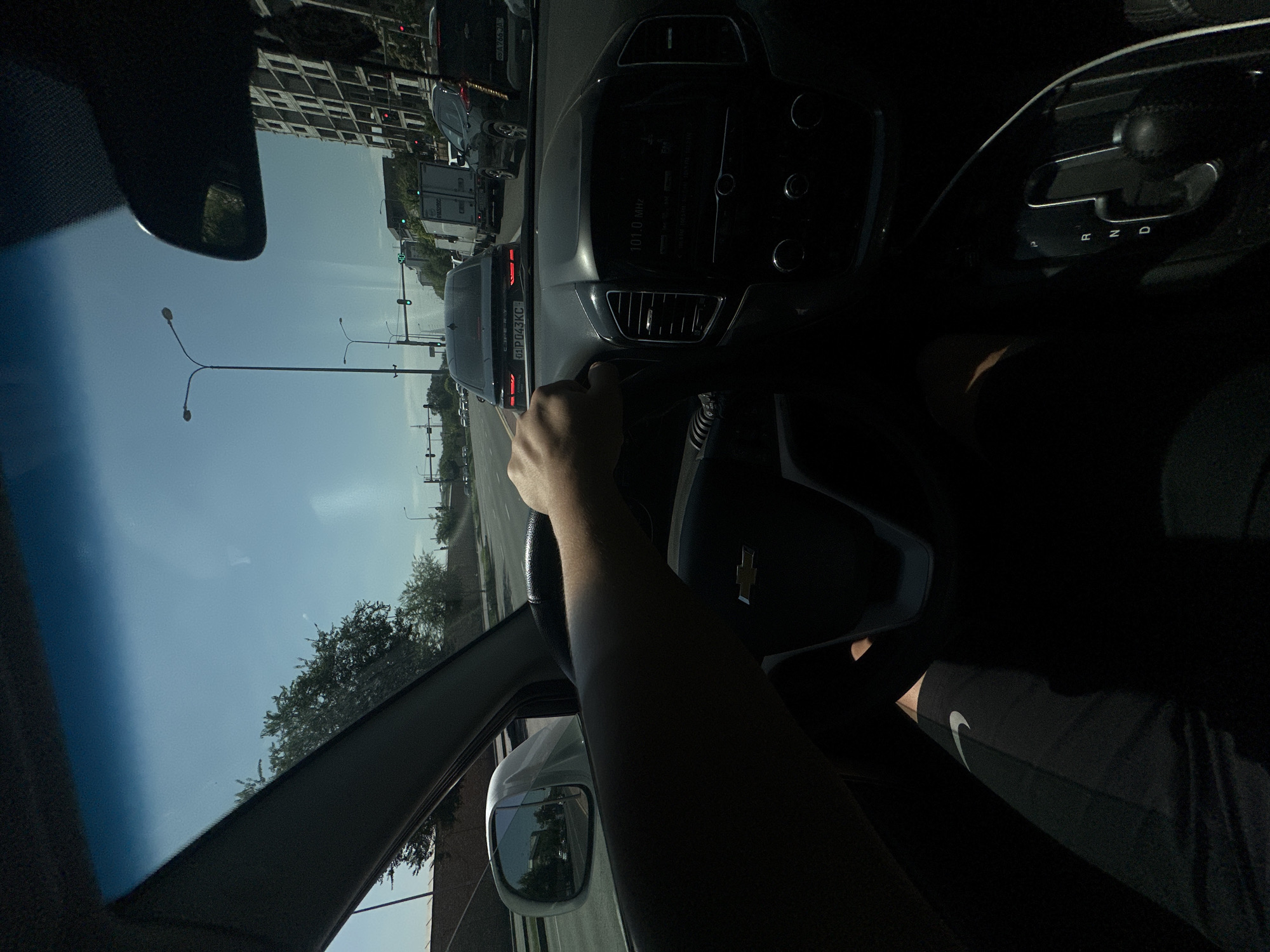 The image shows a first-person perspective from inside a car driving in Tashkent, Uzbekistan. The driver's hand is visible on the steering wheel. The dashboard displays "101.0 MHz" on the radio. Outside the car, traffic is visible, including a black Chery car with a license plate ending in "01 P043KC", a white truck, and other vehicles. There are traffic lights and streetlights, and a tall building under construction is visible on the right side of the image. The sky is clear and blue, indicating daytime. The interior of the car is mostly in shadow, with the driver's legs in dark pants with a Nike logo visible on the left leg.