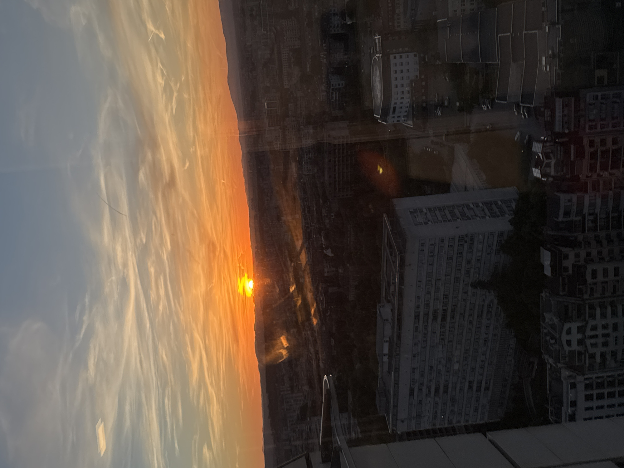 This photo captures a sunset over what appears to be the city of Sofia, Bulgaria. The sky is a gradient of light blue to fiery orange, with whispy clouds stretching across the horizon. The sun is close to the horizon, partially obscured by distant land. Below the horizon is an urban landscape composed of buildings with varying heights and architectural styles. The photo is taken from a high vantage point, possibly from a skyscraper or high-rise building, evident by the railing or ledge visible in the foreground. The scene has a reflective quality, perhaps captured through glass or a window, with the urban landscape overlayed on the sunset sky.