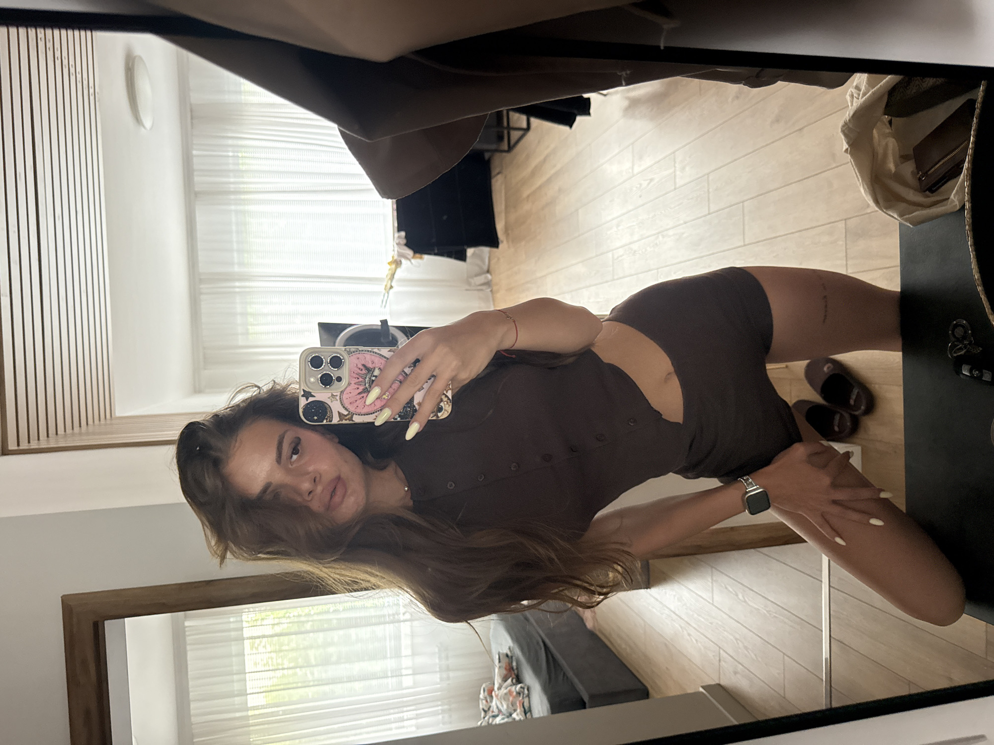 This is a vertical, full-body mirror selfie of a young woman in Plovdiv, Bulgaria. She is wearing a brown, short-sleeved romper with a button-down front and cutouts on the sides. Her hair is long and brown, and she has light makeup on. She is holding a smartphone in her right hand, which has a pink phone case with a floral design. On her left wrist, she wears a silver smartwatch.
The setting appears to be an indoor space, likely a bedroom or dressing area, with light-colored wooden floors and white walls. To the left of the woman is a light wood slatted panel and a white circular light fixture on the wall. In the background, sheer white curtains cover a window, allowing natural light to filter in. The reflection in the mirror also shows a dark couch with a patterned throw blanket on it in the far background.
The woman is posing with her left leg extended and her right arm resting on her thigh. Her expression is relaxed and slightly playful. The lighting suggests it is daytime. There is no visible text in the image.